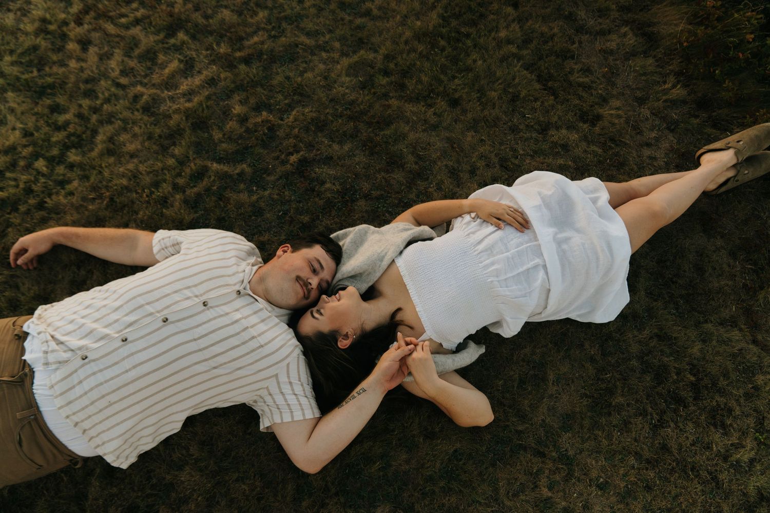 Couple lying on grass holding hands, wearing white outfits, viewed from above in a romantic outdoor setting.