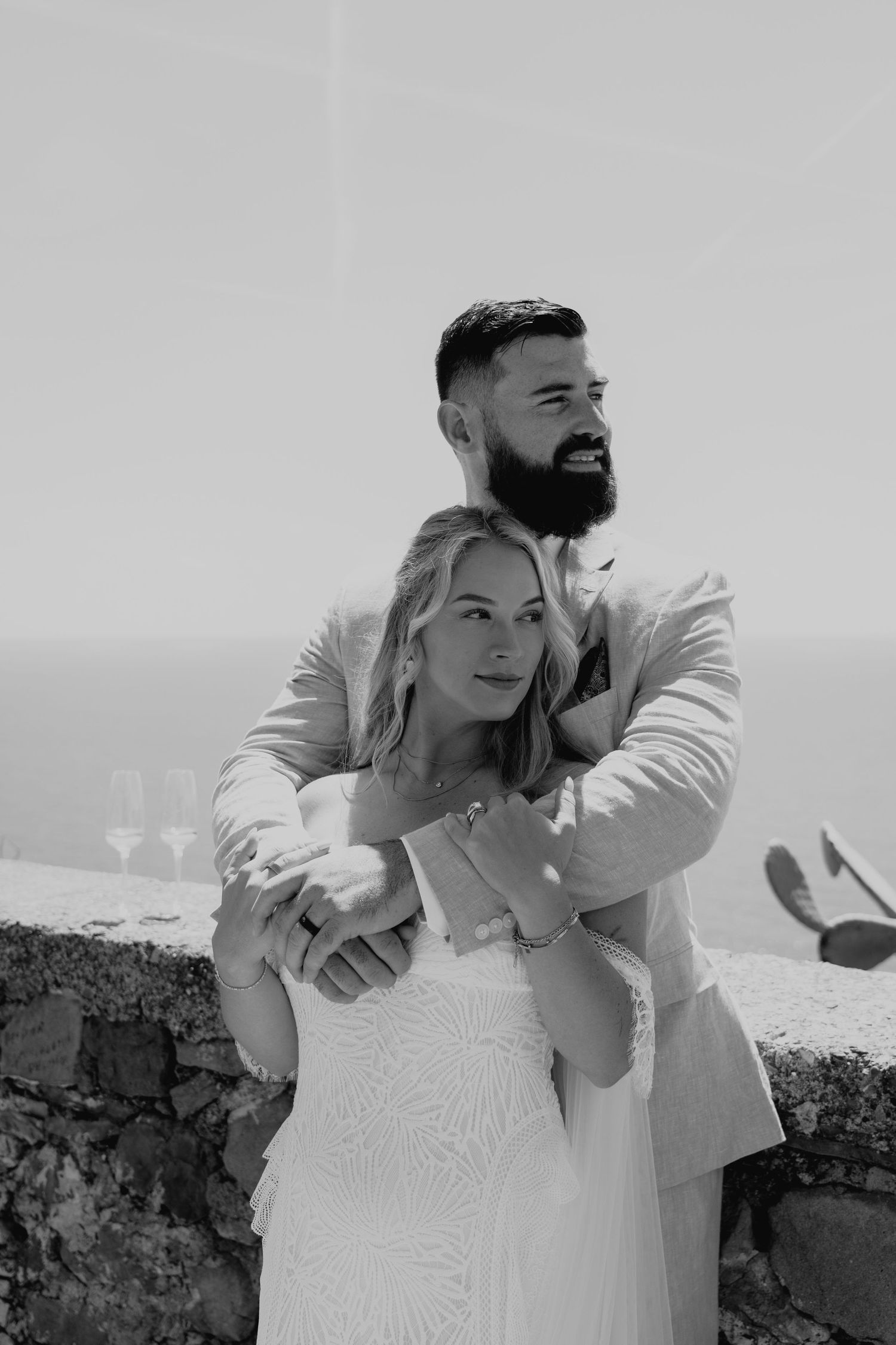 A black and white wedding photo series shows a couple embracing and laughing together against a stone wall backdrop.