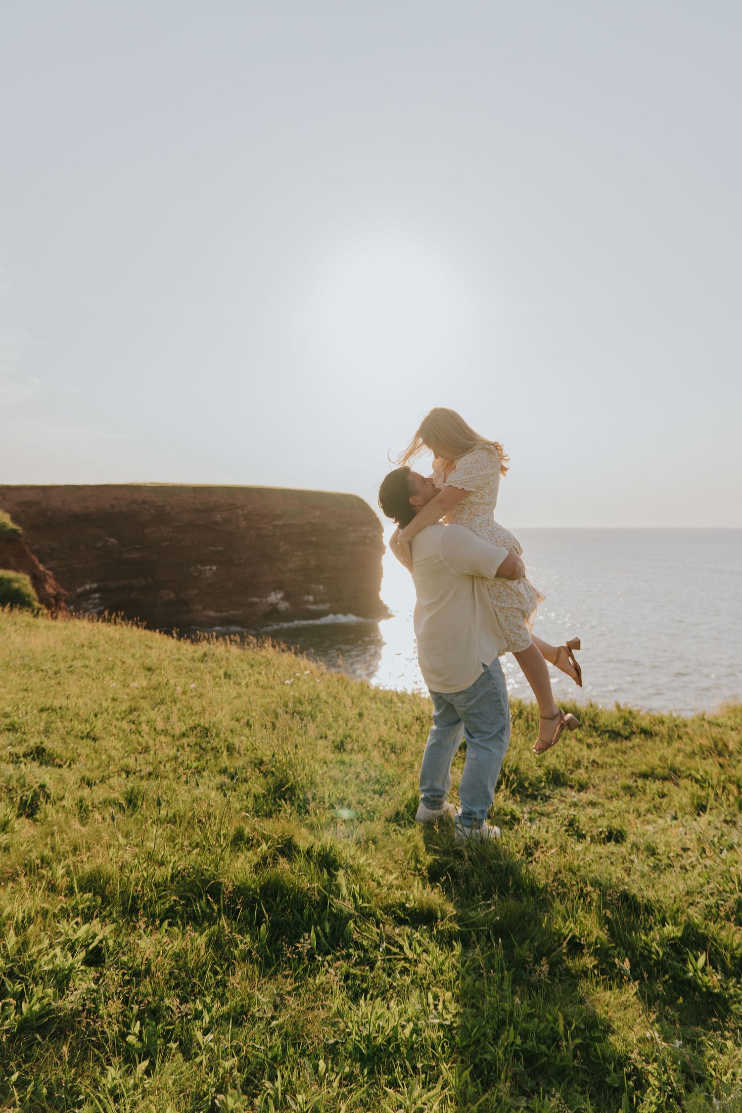 A person in a white sweater walks along a grassy coastal cliff edge at sunset, creating a dreamy silhouette effect.