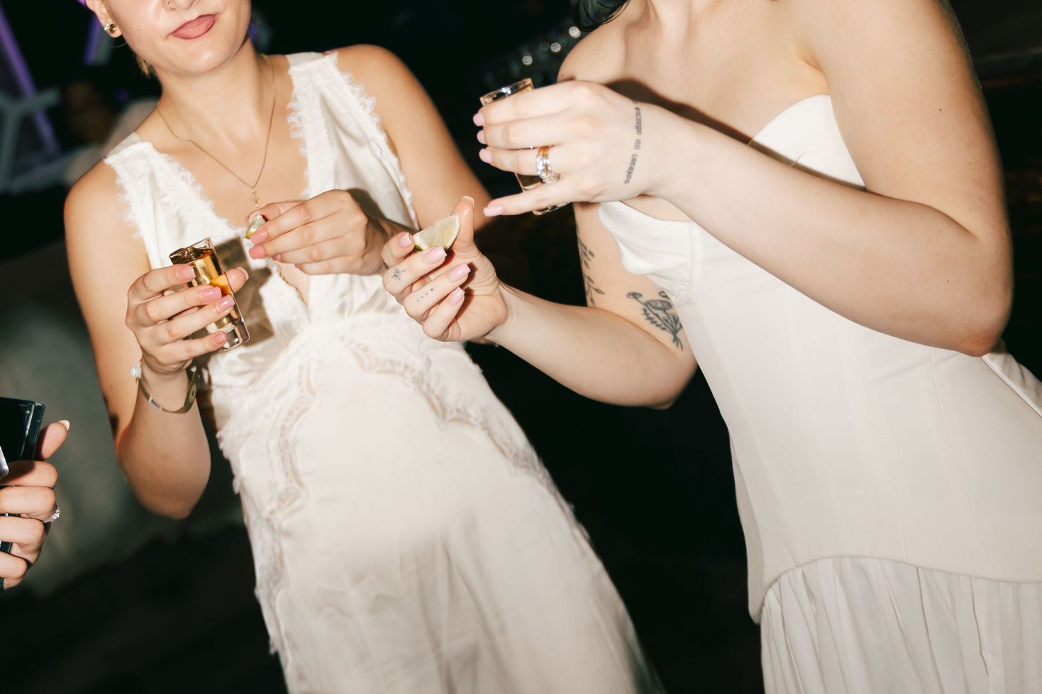 Two women in white dresses holding drinks and sharing a moment at a wedding reception.