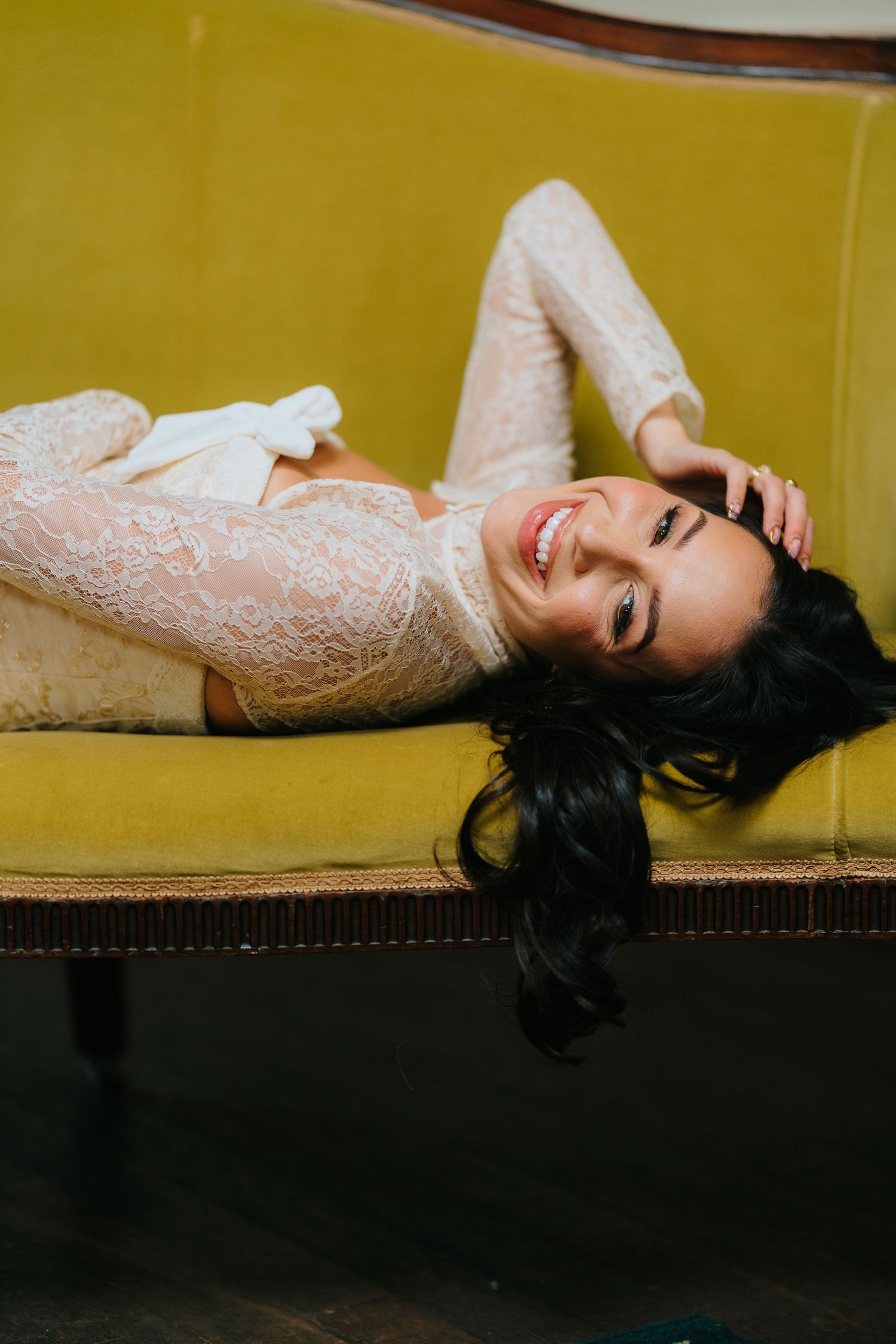 Smiling bride in white lace gown lying on a vintage yellow velvet sofa, looking up with dark hair spread out.