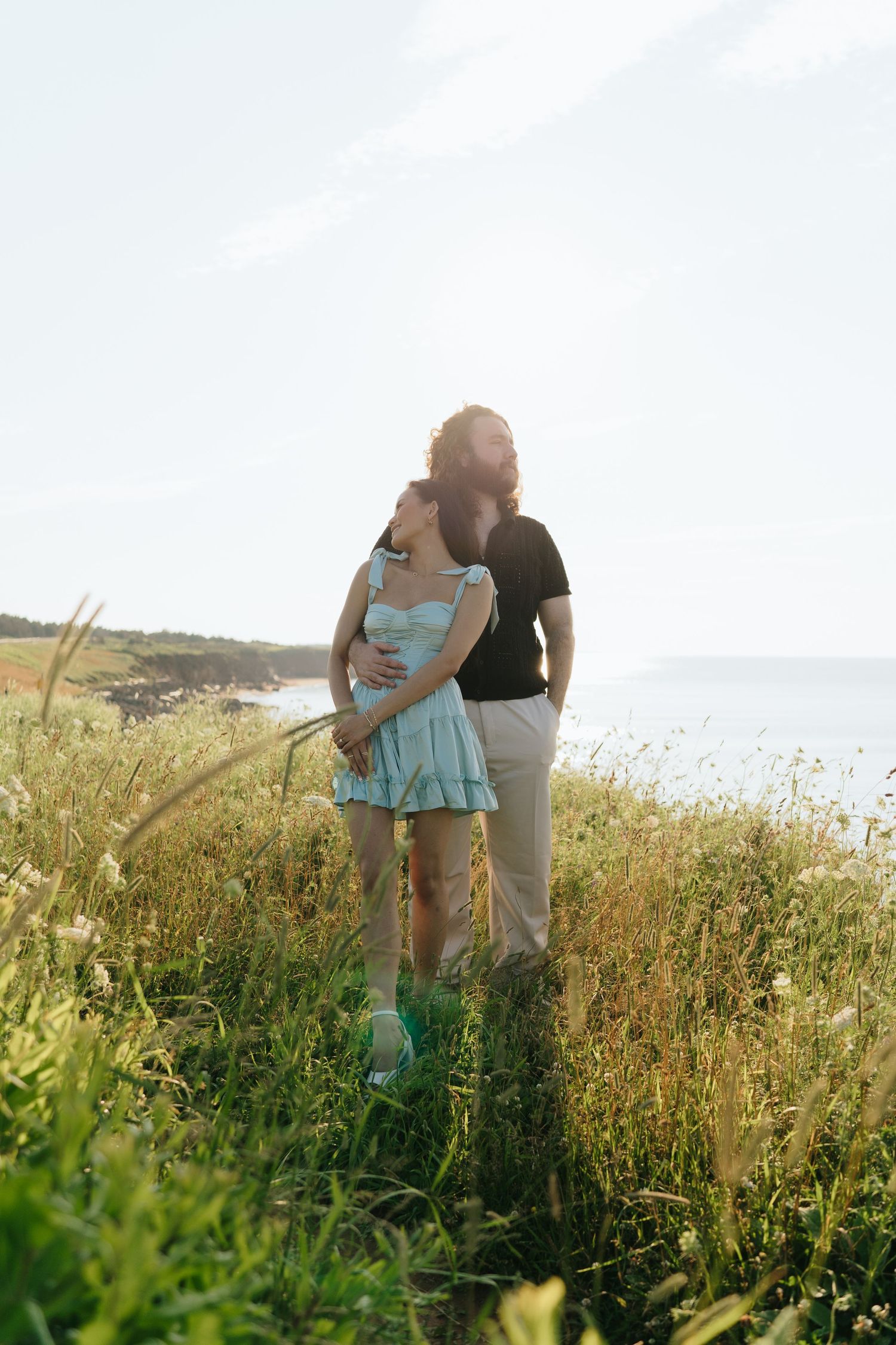 A romantic couple stands together in a field of tall grass near the ocean.