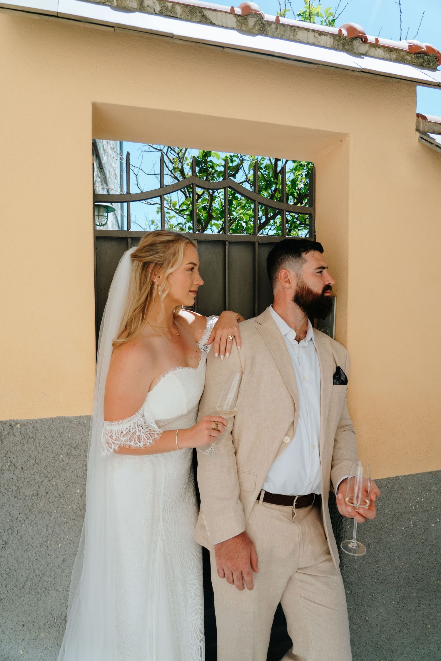 A couple in wedding attire share a playful moment against a beige wall with windows.