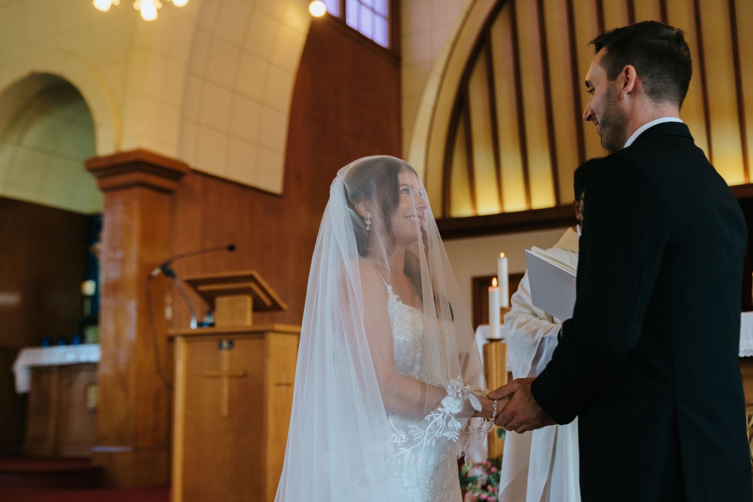Bride in white gown and veil exchanging vows with groom in black suit at a church wedding ceremony altar.