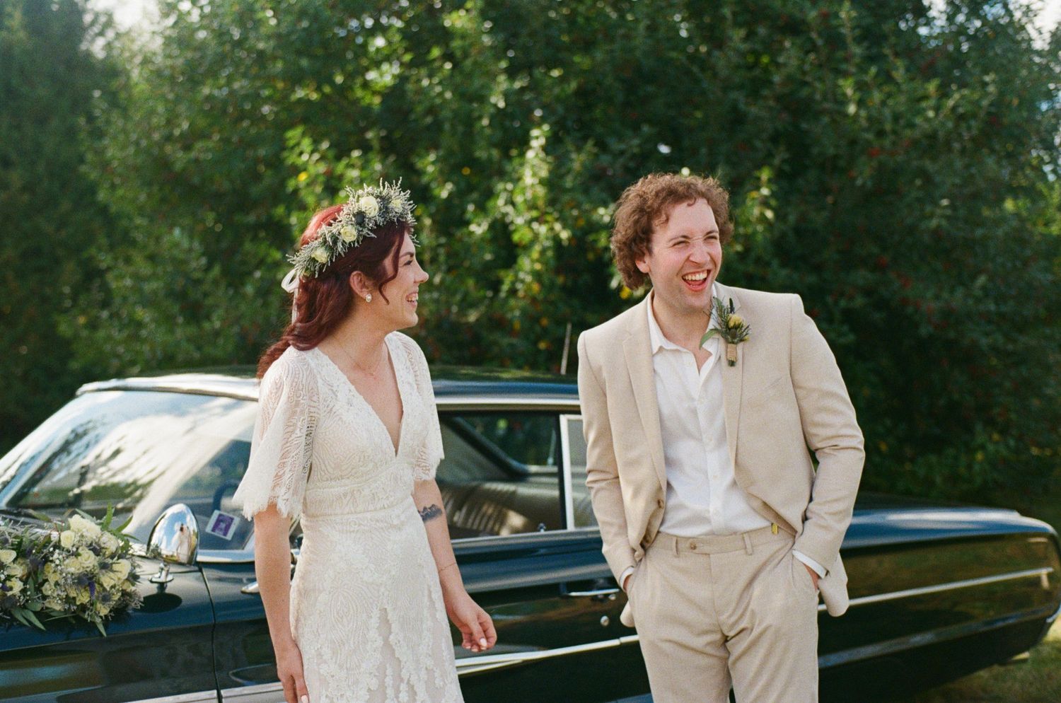 Newlyweds in light-colored attire standing by vintage car in summer garden setting.