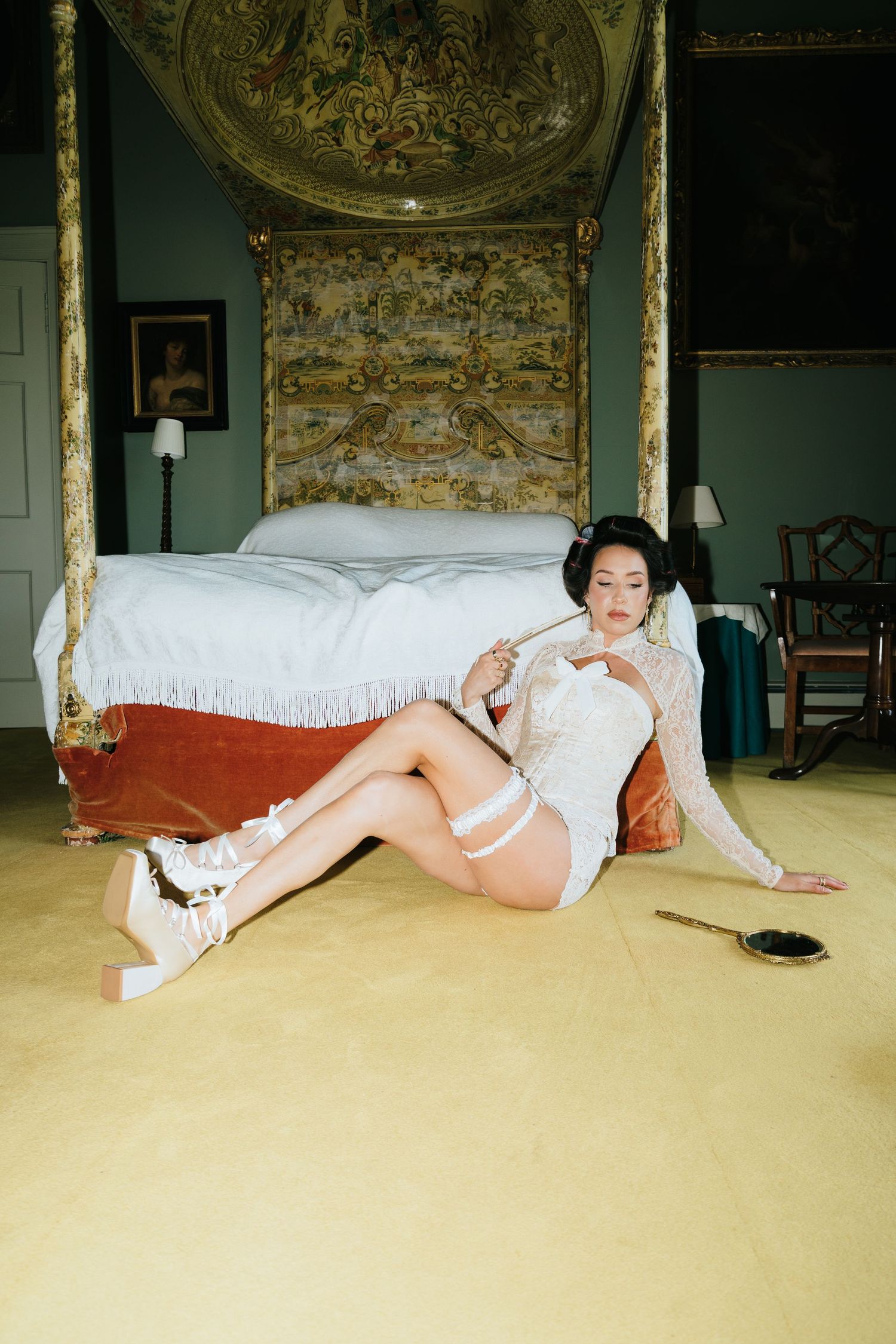 Woman in white lace lingerie reclining on the floor beside an antique four-poster bed in an elegant room.