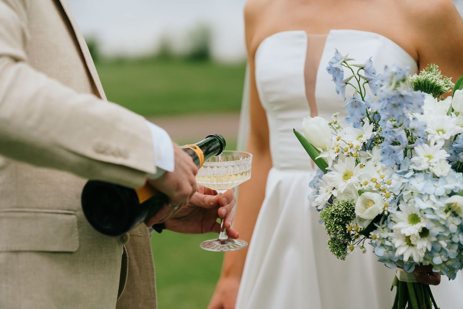 Groom in beige suit pouring champagne for bride holding blue hydrangea bouquet at outdoor wedding celebration.