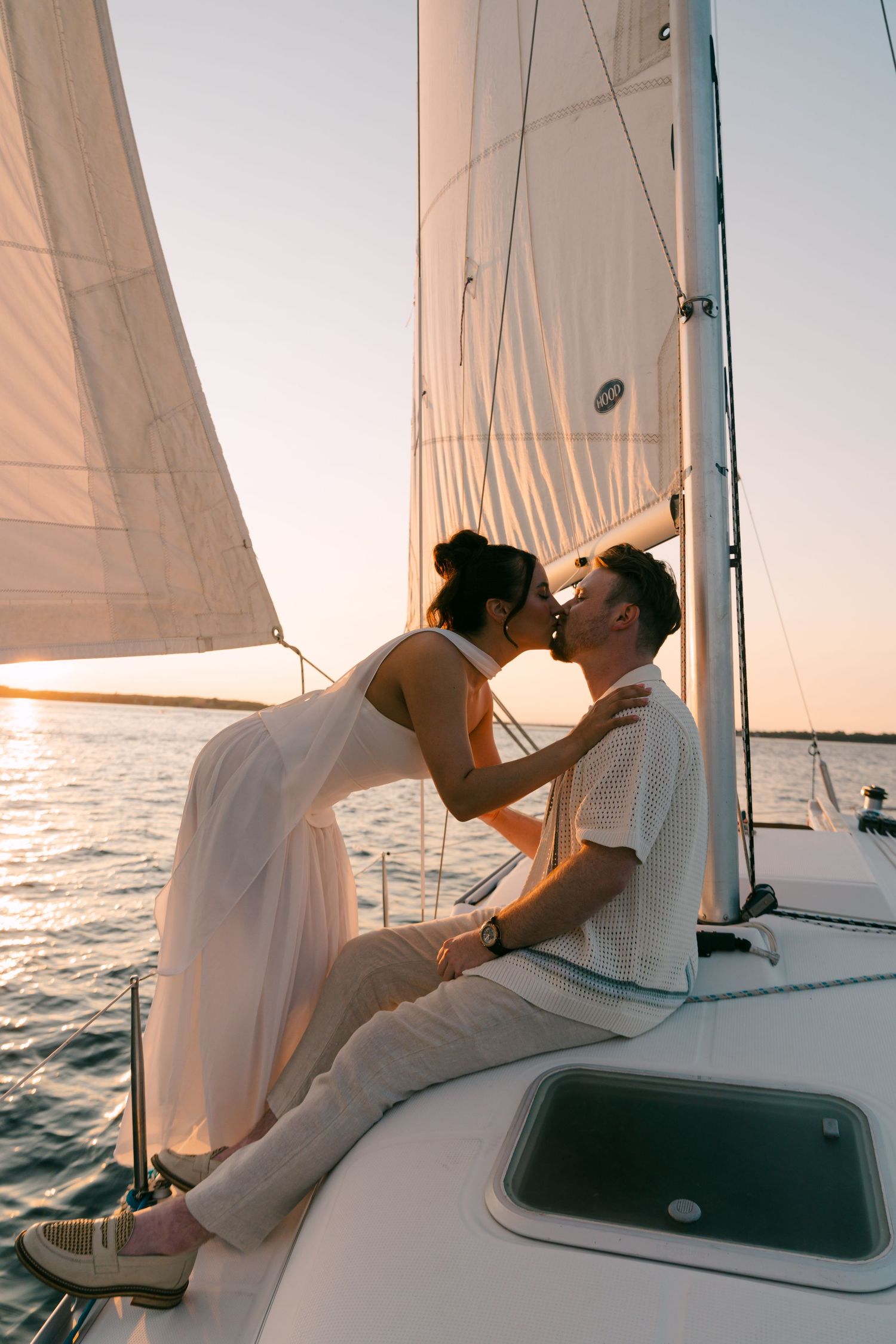 Romantic couple kissing on a sailboat at sunset, woman in white dress, golden hour light reflecting on calm water.