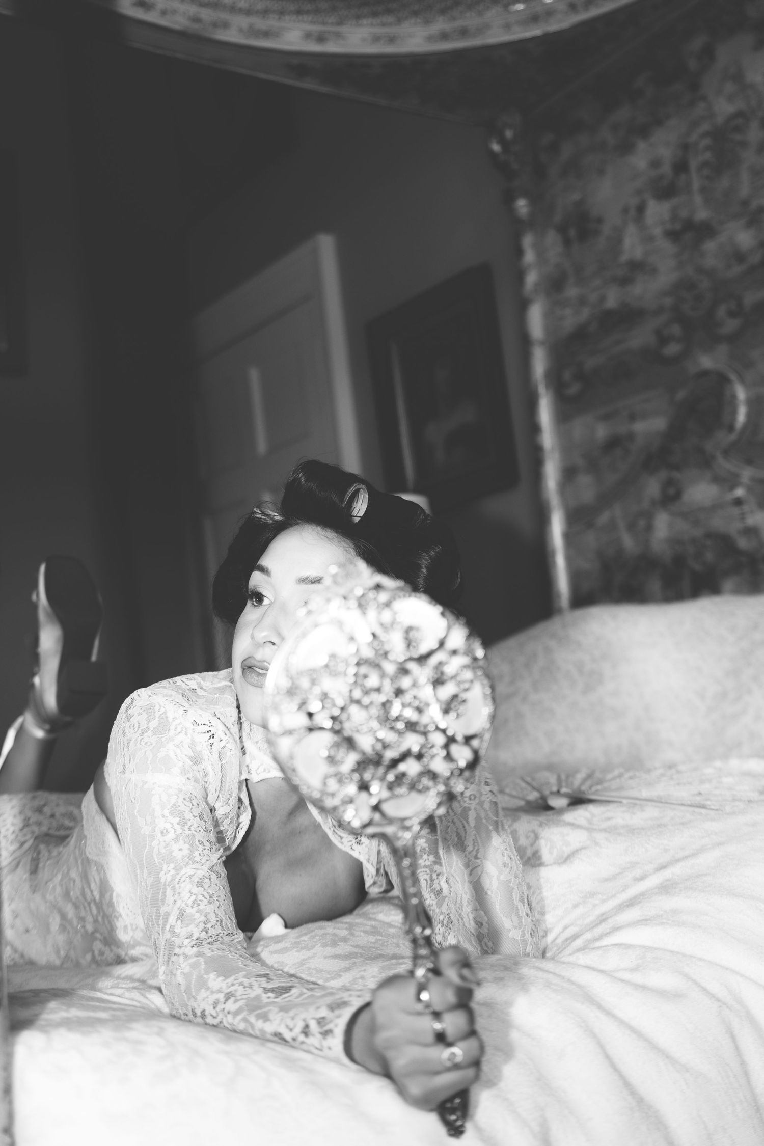 Black and white photo of a bride lying on a bed holding a floral bouquet, wearing lace dress with hair rollers.