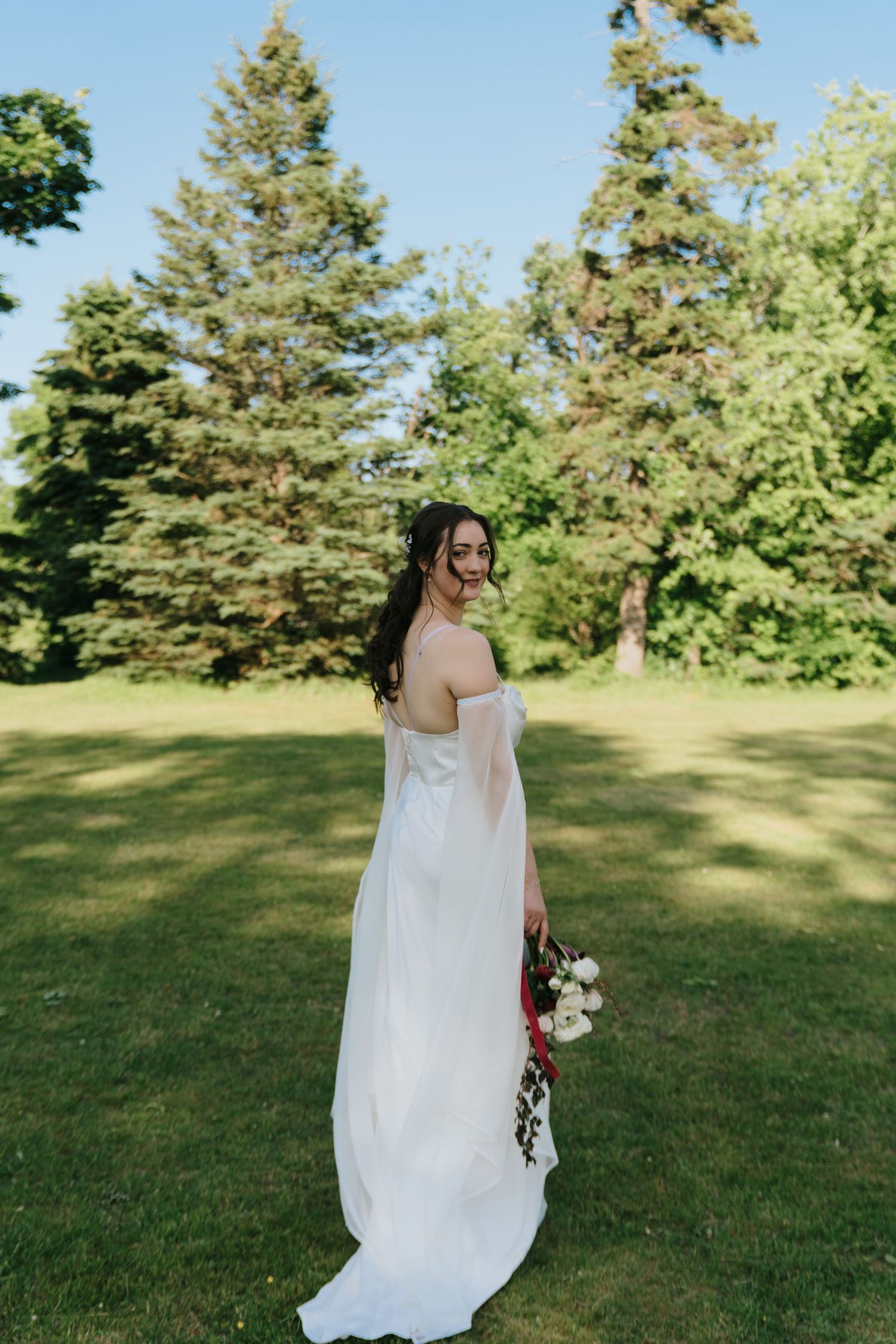 A bride in a flowing white dress stands in a sunny field surrounded by evergreen trees.