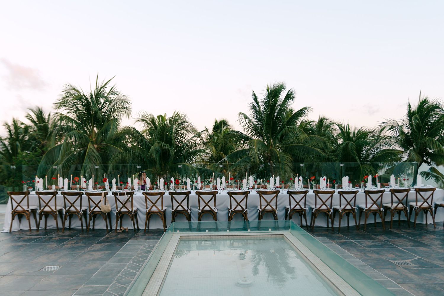 Elegant outdoor wedding reception with long dining table, wooden chairs, white linens, and tropical palm trees by a pool.