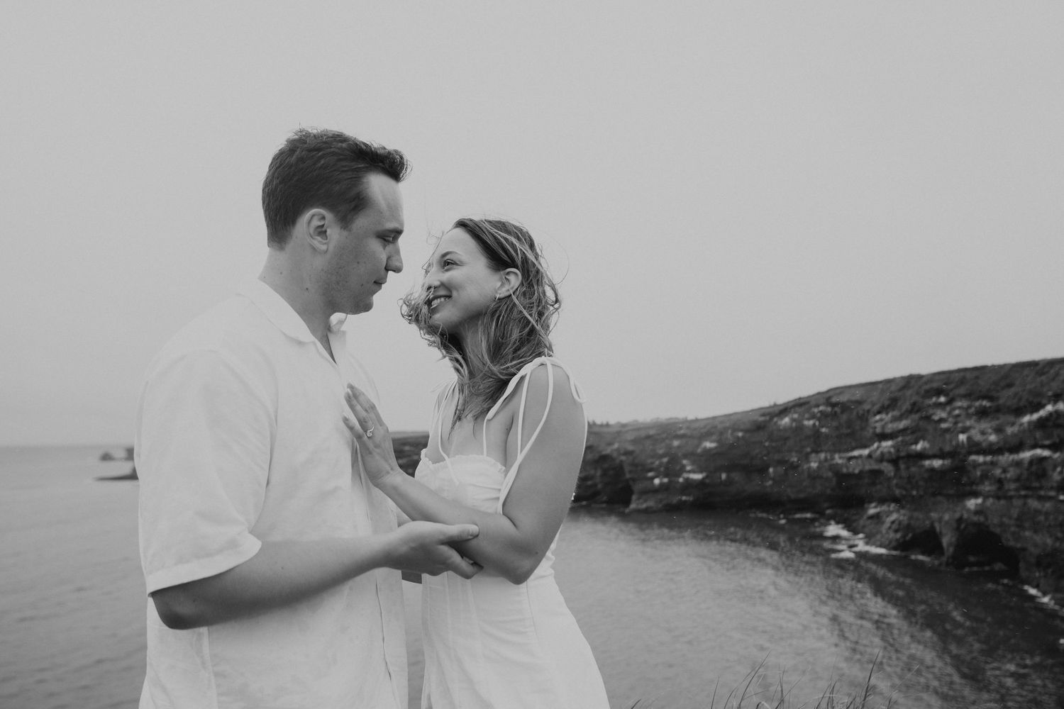 A romantic black and white photo series of a couple embracing on a rocky coastal landscape.