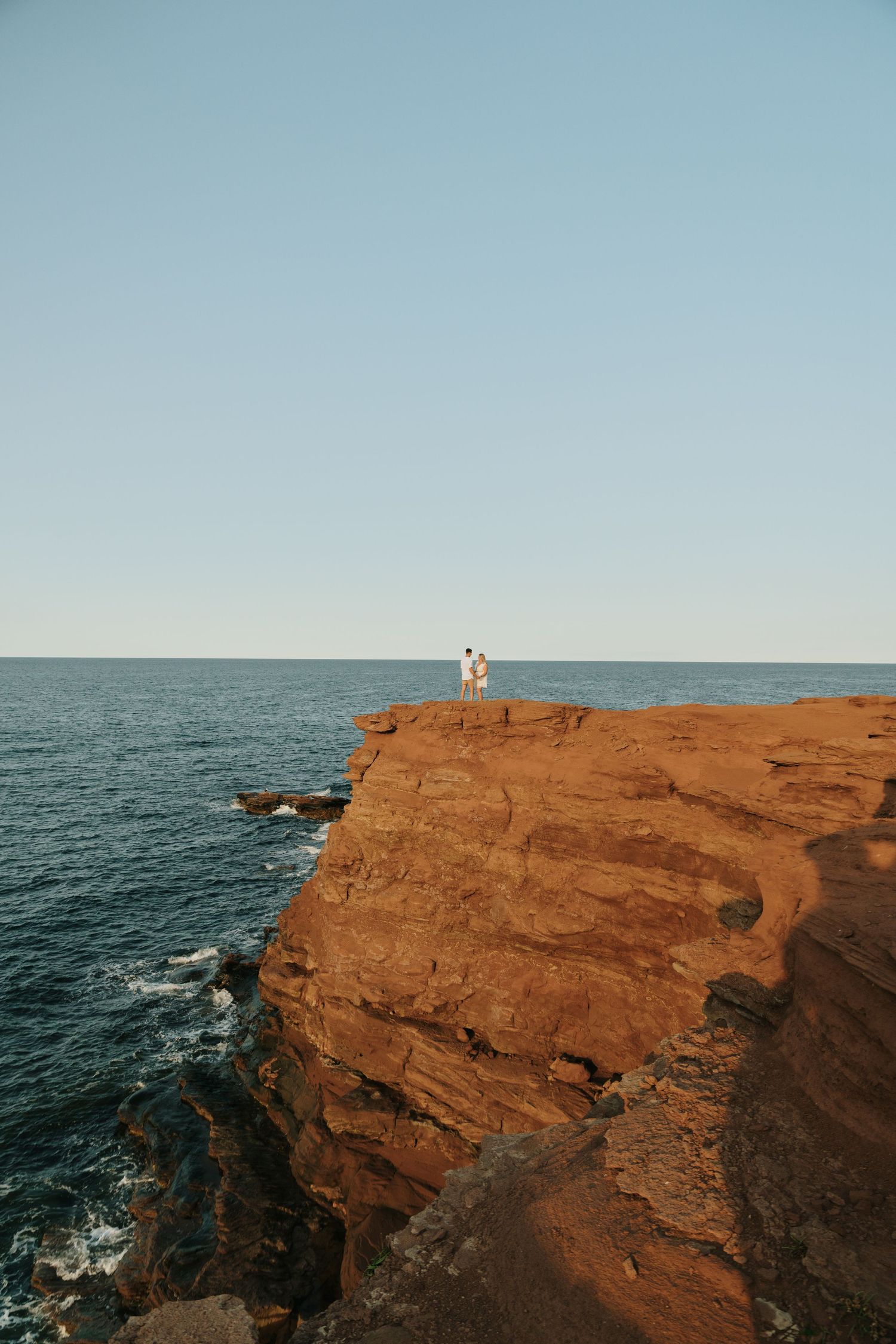 A person stands on the edge of dramatic red cliffs overlooking the blue ocean waters at sunset in Cape Tryon, PEI