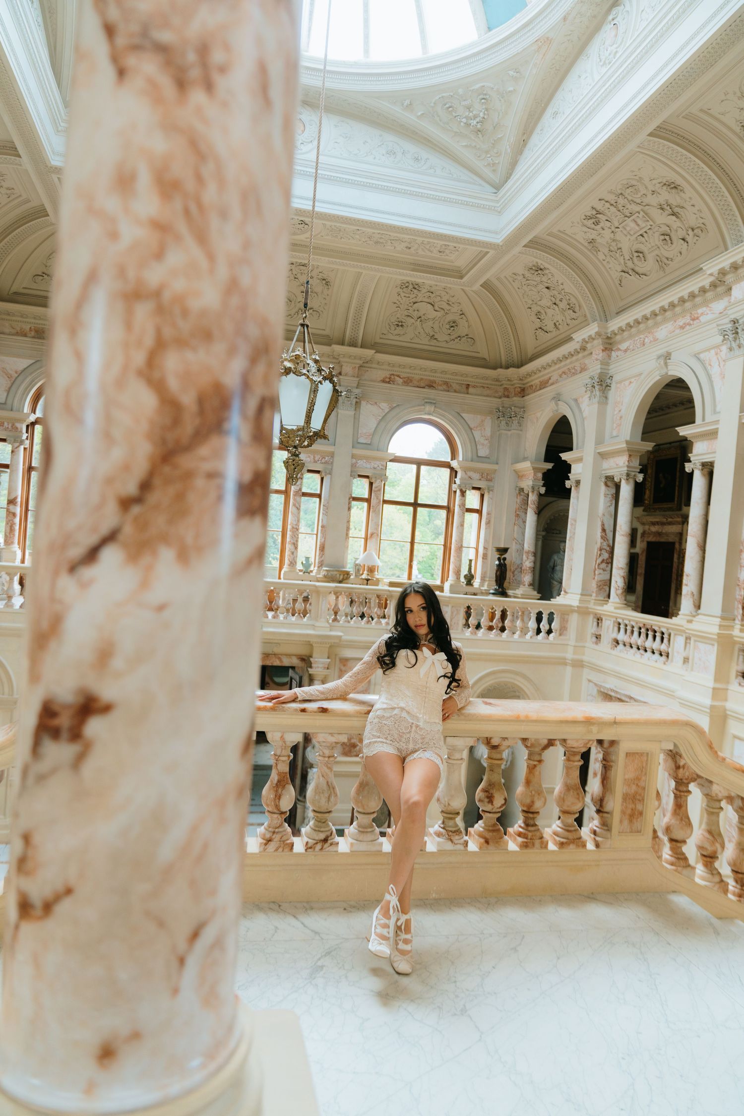 Woman in white shorts poses by ornate marble balustrade in a luxurious classical palace interior.