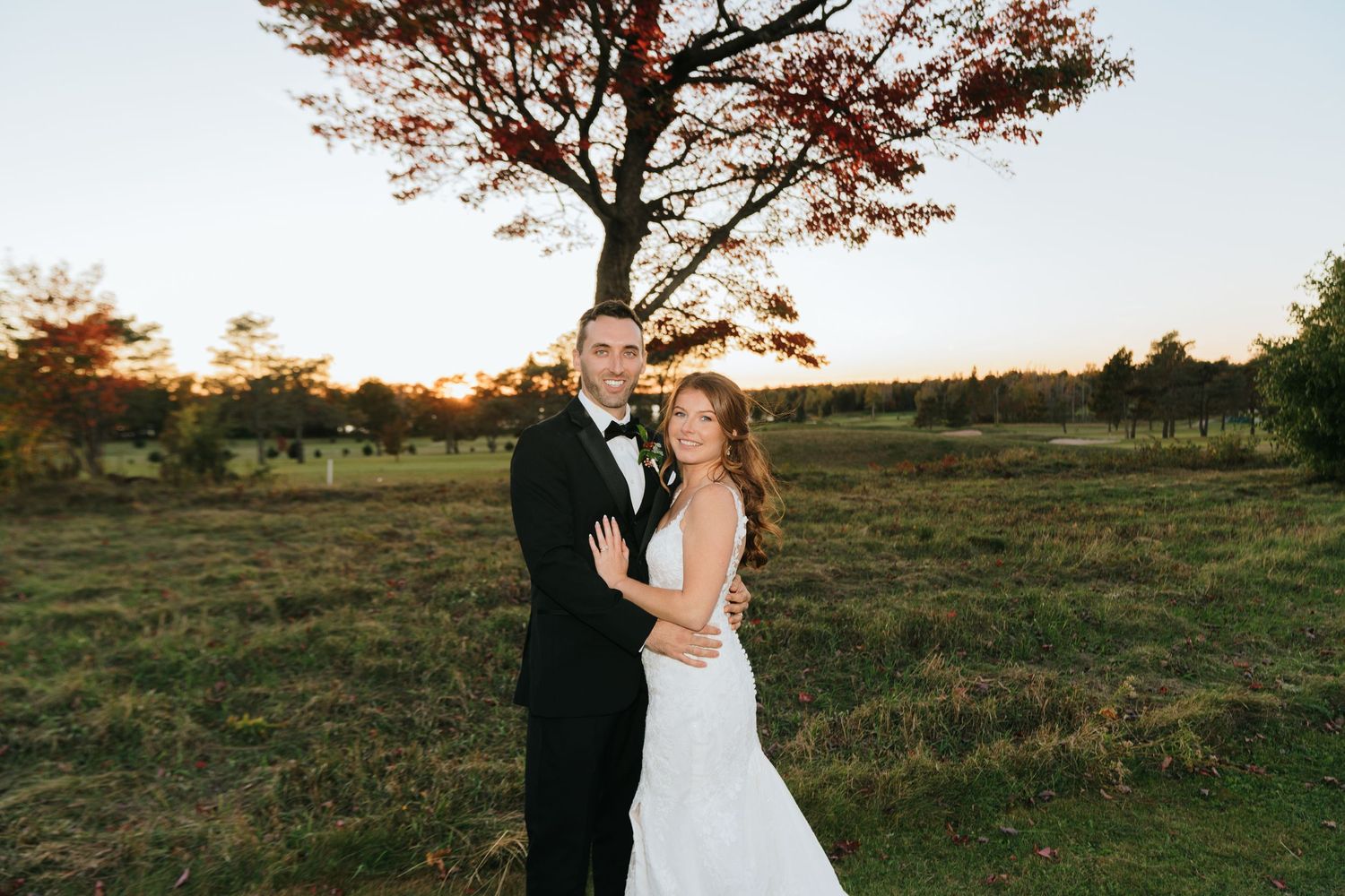 Bride and groom pose together at golden hour near a tree in an open field, wearing wedding gown and black tuxedo.