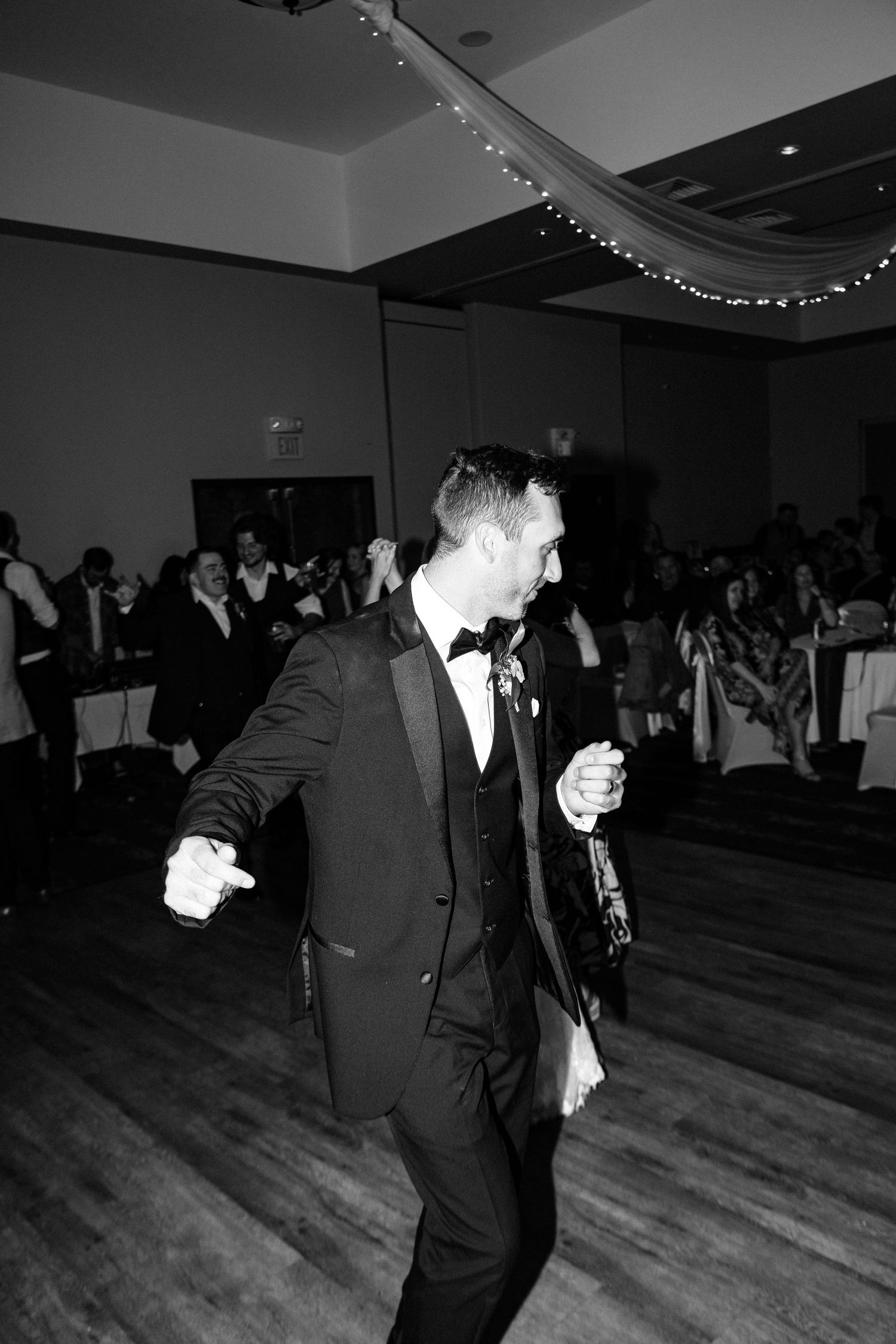 Groom in black tuxedo dancing joyfully at wedding reception in elegant ballroom, black and white photo.