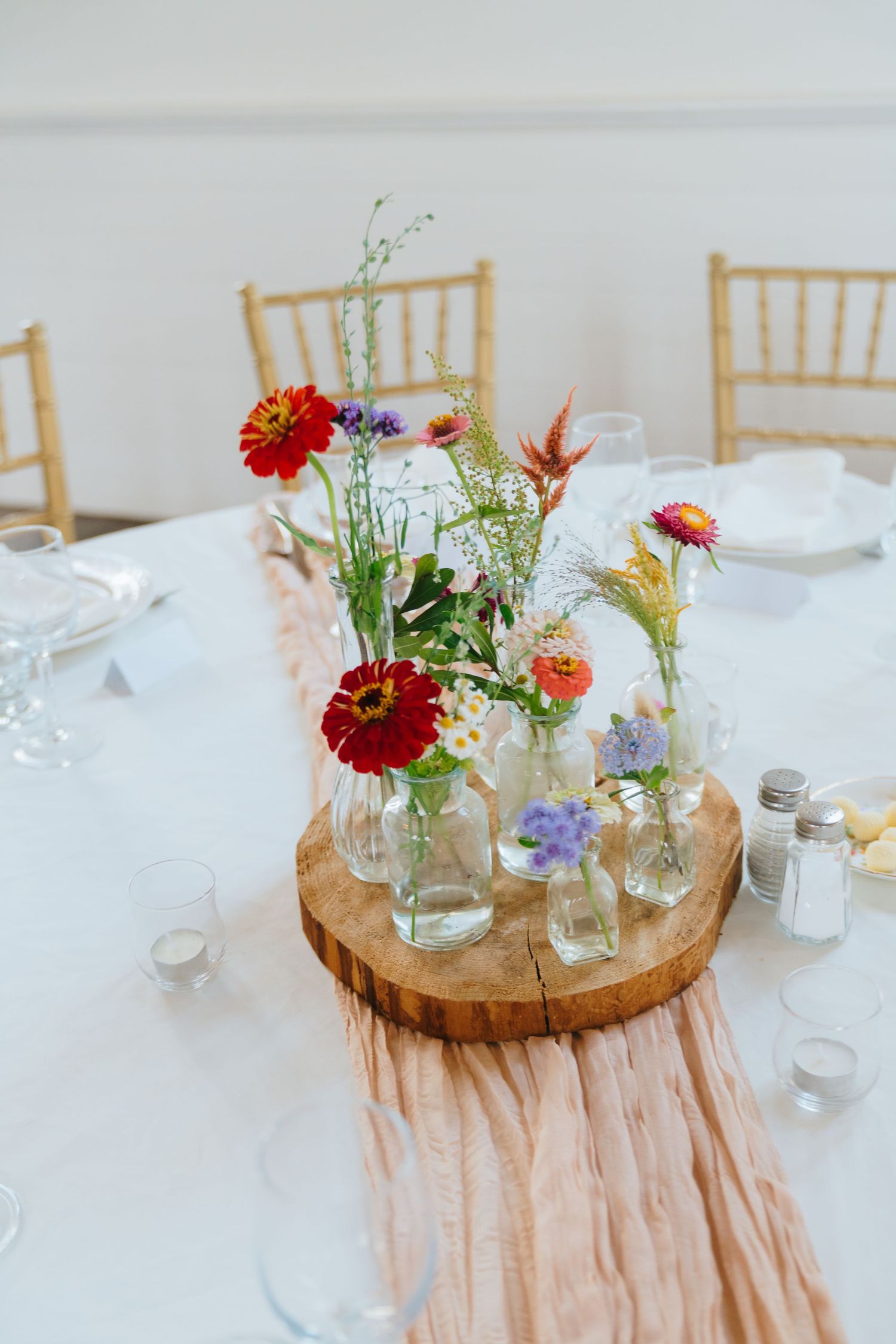 Rustic wedding centerpiece with red zinnias and wildflowers in glass bottles on a wooden slice.