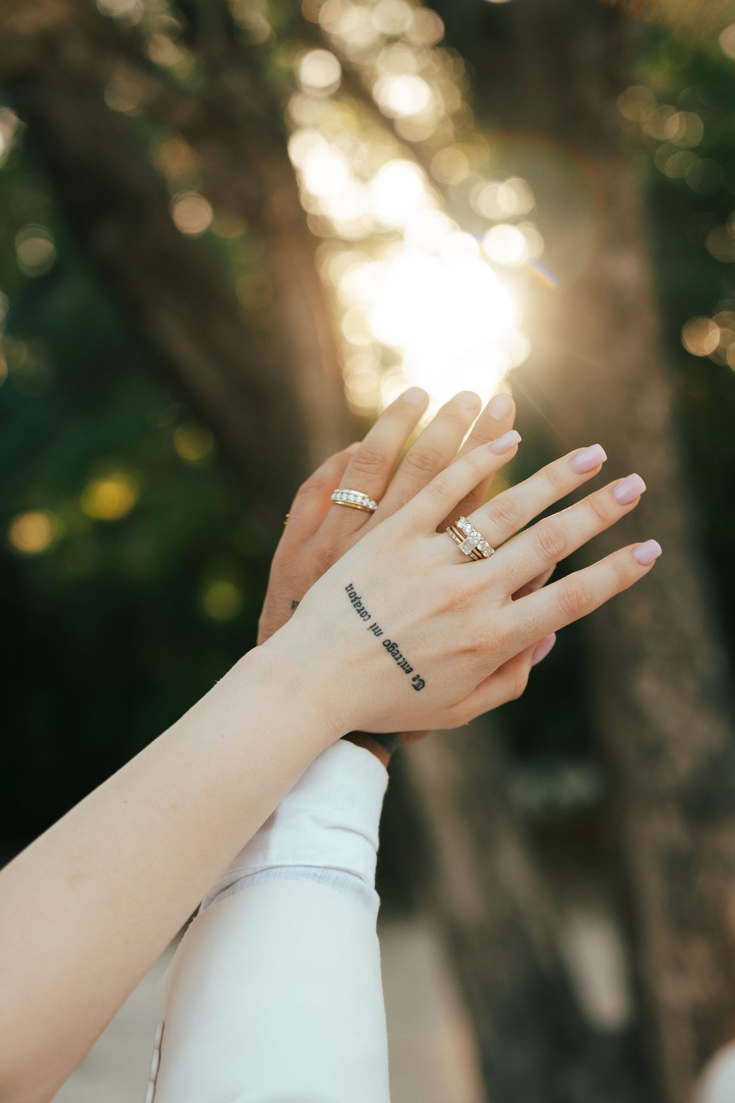 Two brides display wedding rings and a tattoo, hands raised against sunlit trees.