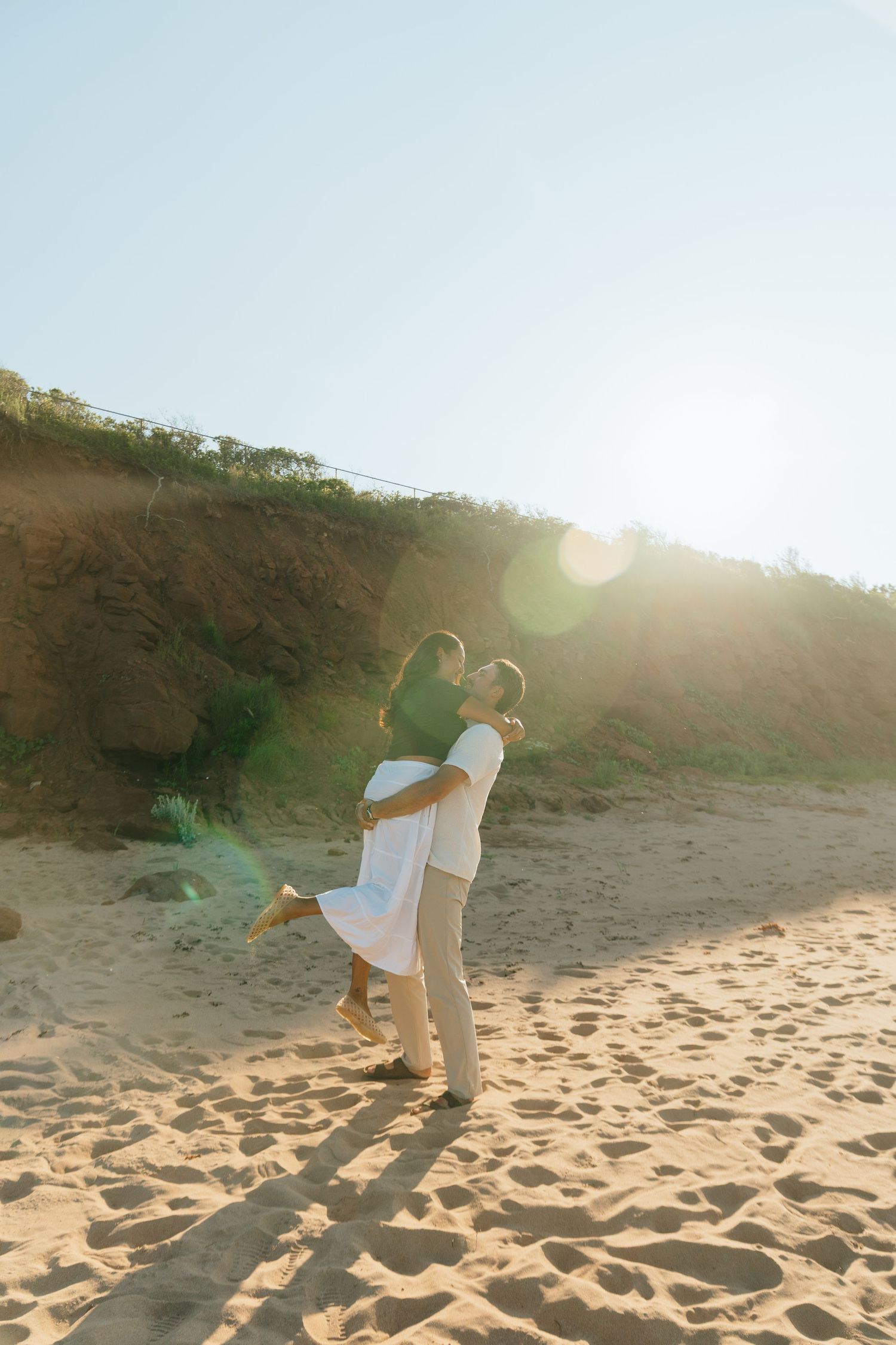 Couple embracing on a sunny sandy beach, man lifting woman in white dress with golden dunes and bright sunlight behind them.