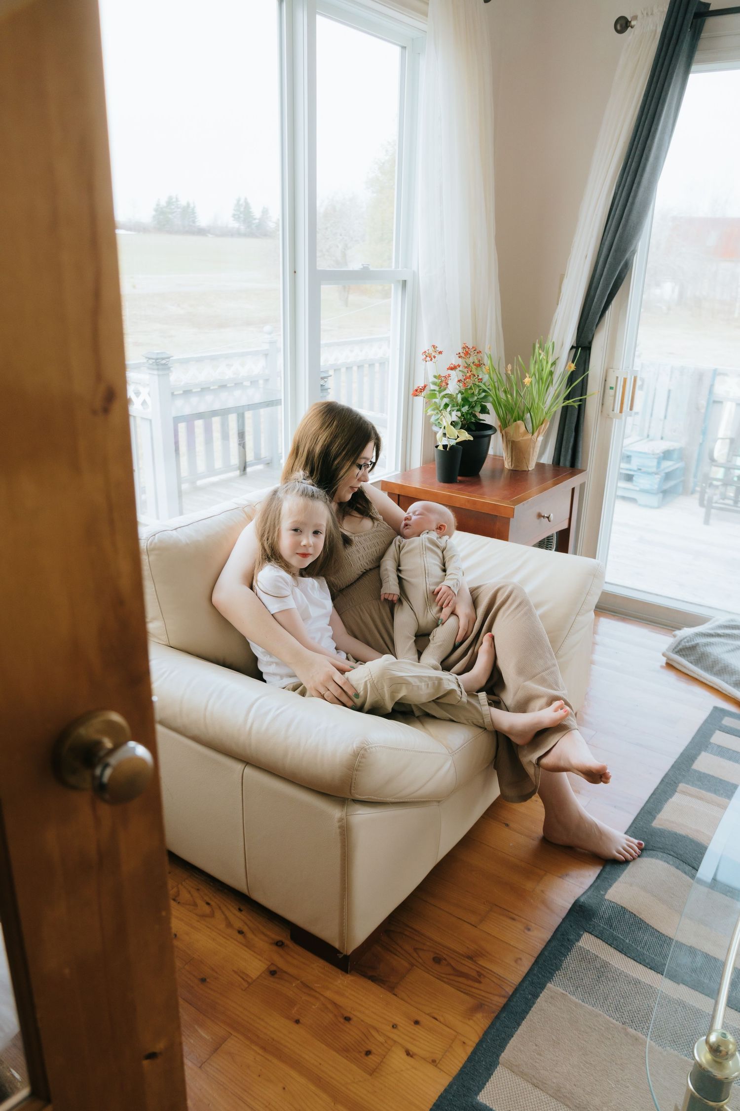 Mother sitting on white sofa with young child and newborn baby in cozy living room with large windows.