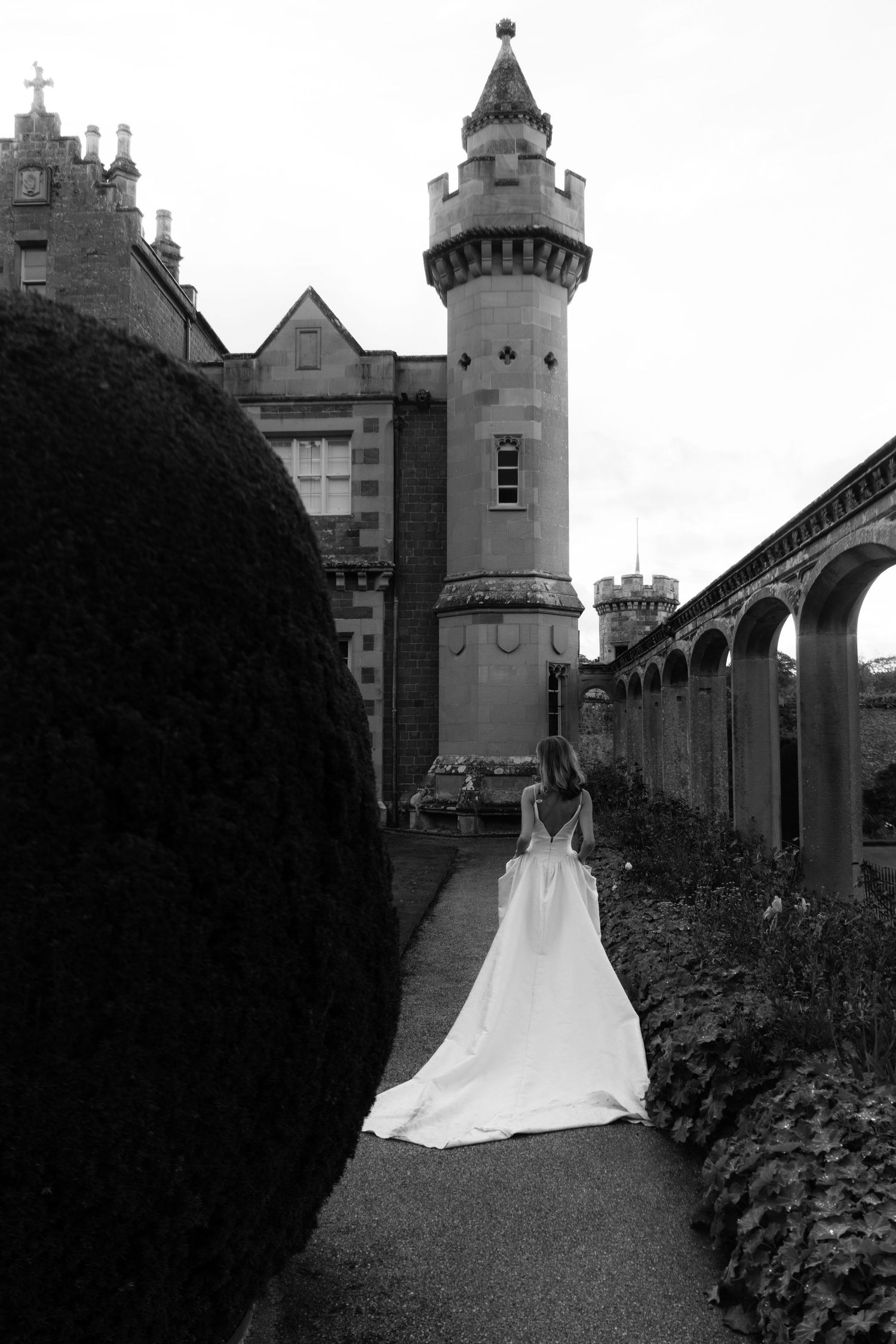 Black and white photo of a bride in a white gown walking toward a castle tower along a garden path.
