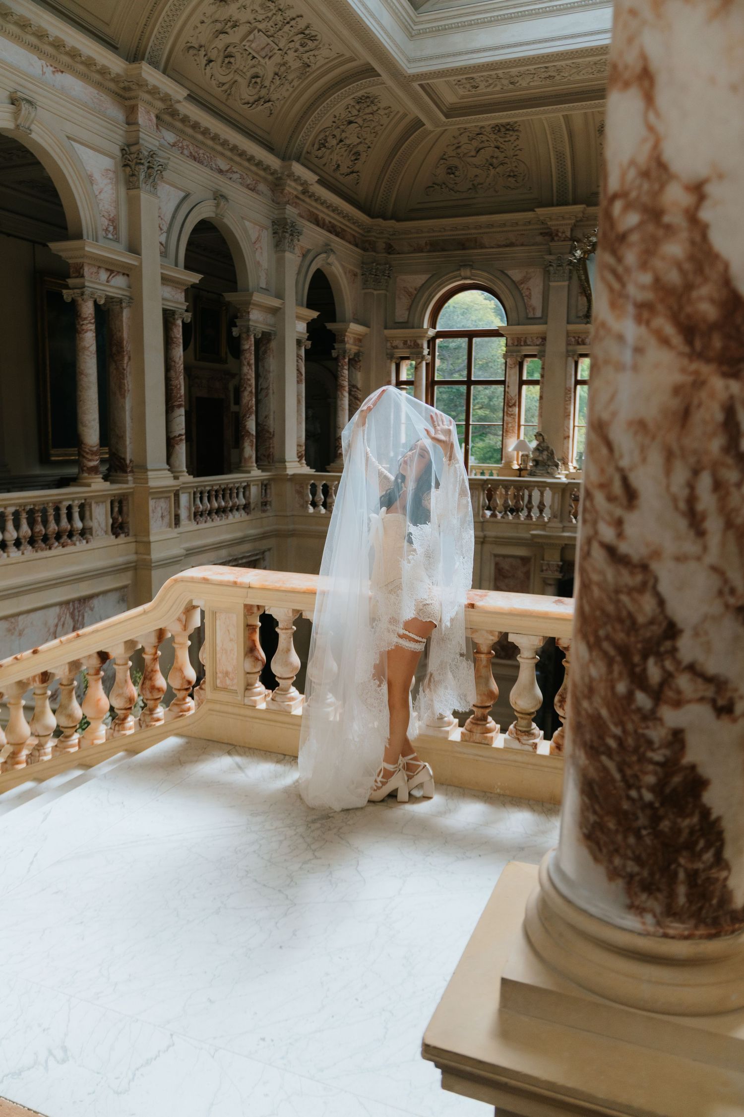 Bride in white wedding gown and veil kissing groom on ornate marble staircase in elegant baroque venue.