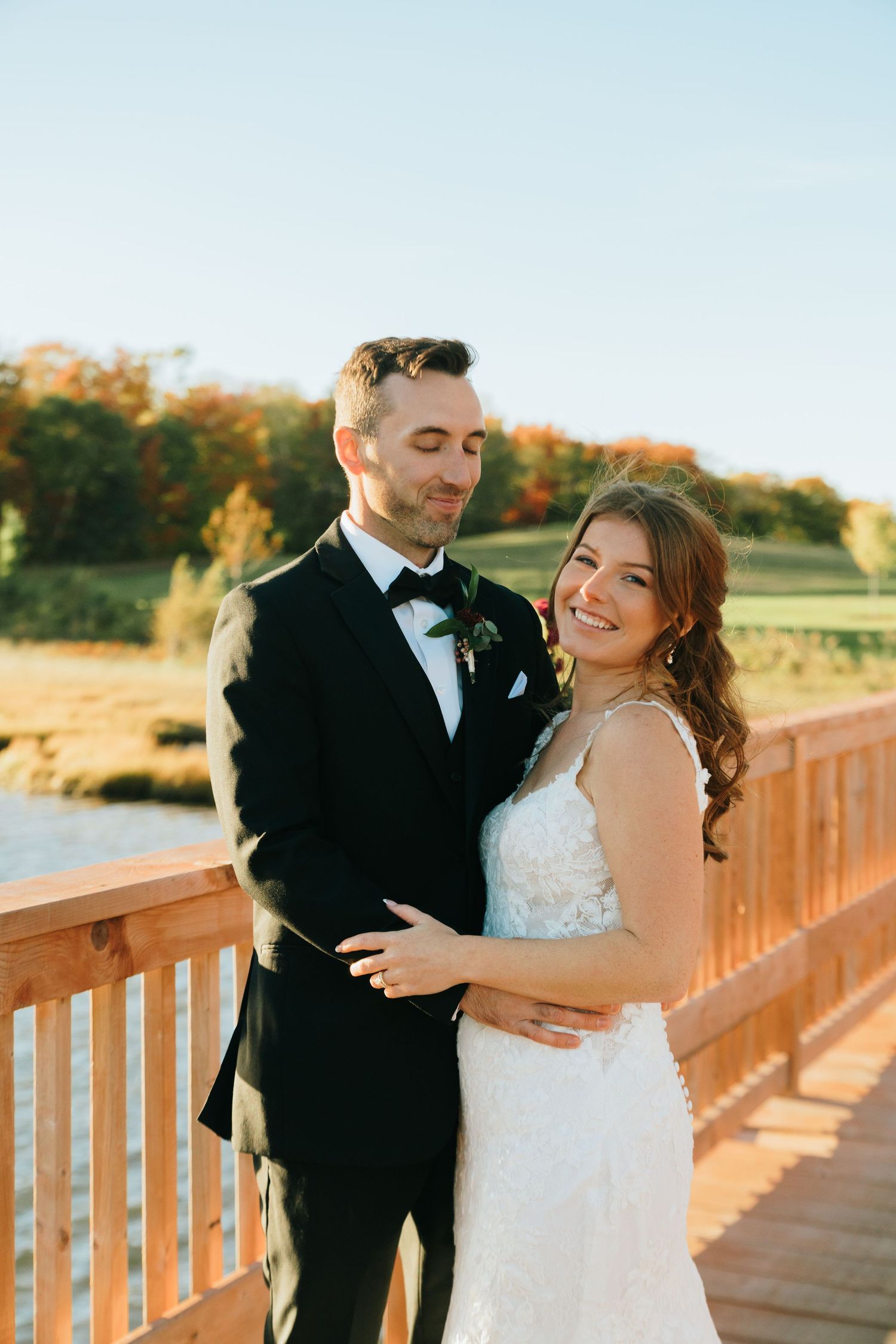 Bride and groom embracing on wooden bridge at sunset with autumn foliage in background.