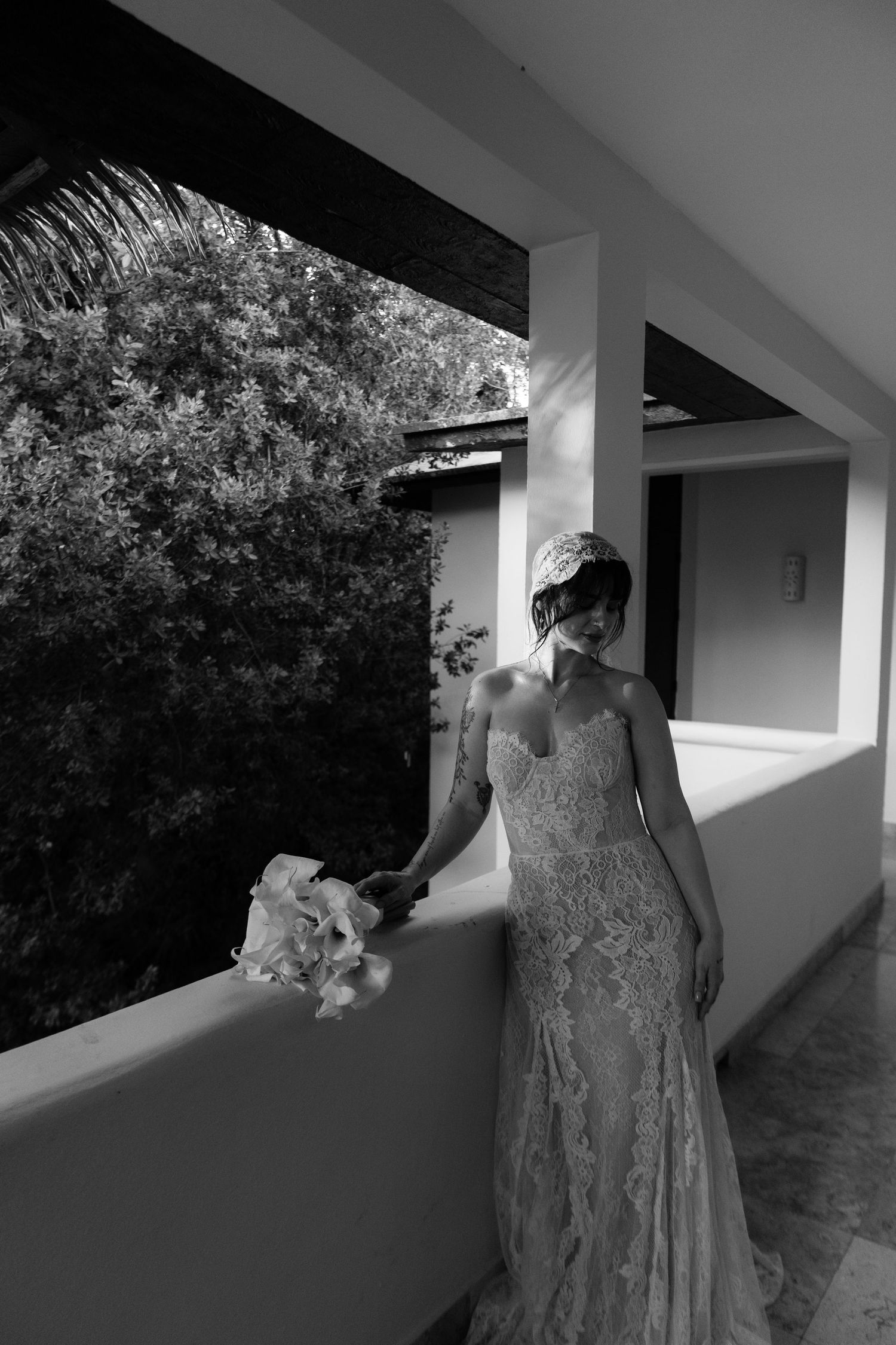 Bride in lace gown holds bouquet on sunlit balcony in black and white photo.
