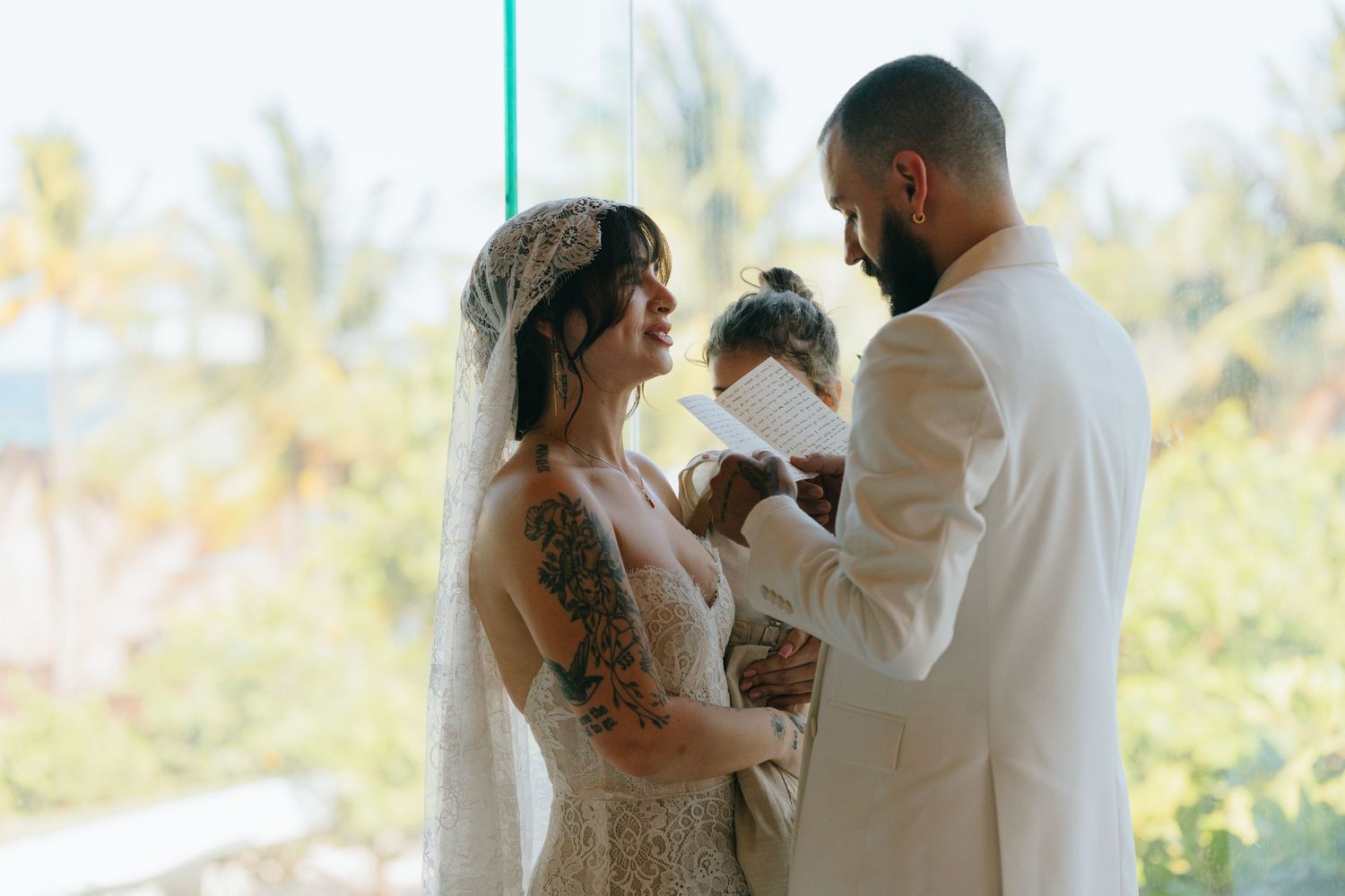 Bride and groom exchange vows at tropical wedding ceremony, bride wearing lace dress with veil and floral tattoos.