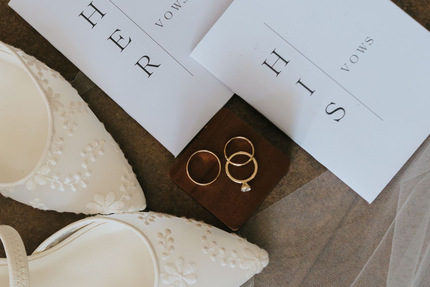 Wedding flat lay with white lace bridal shoes, gold rings on wooden box, and HER and HIS vow booklets.