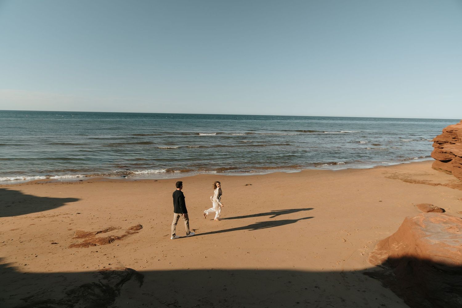 Couple walking hand in hand on a sandy beach with red cliffs and calm blue ocean waves under a clear sky.