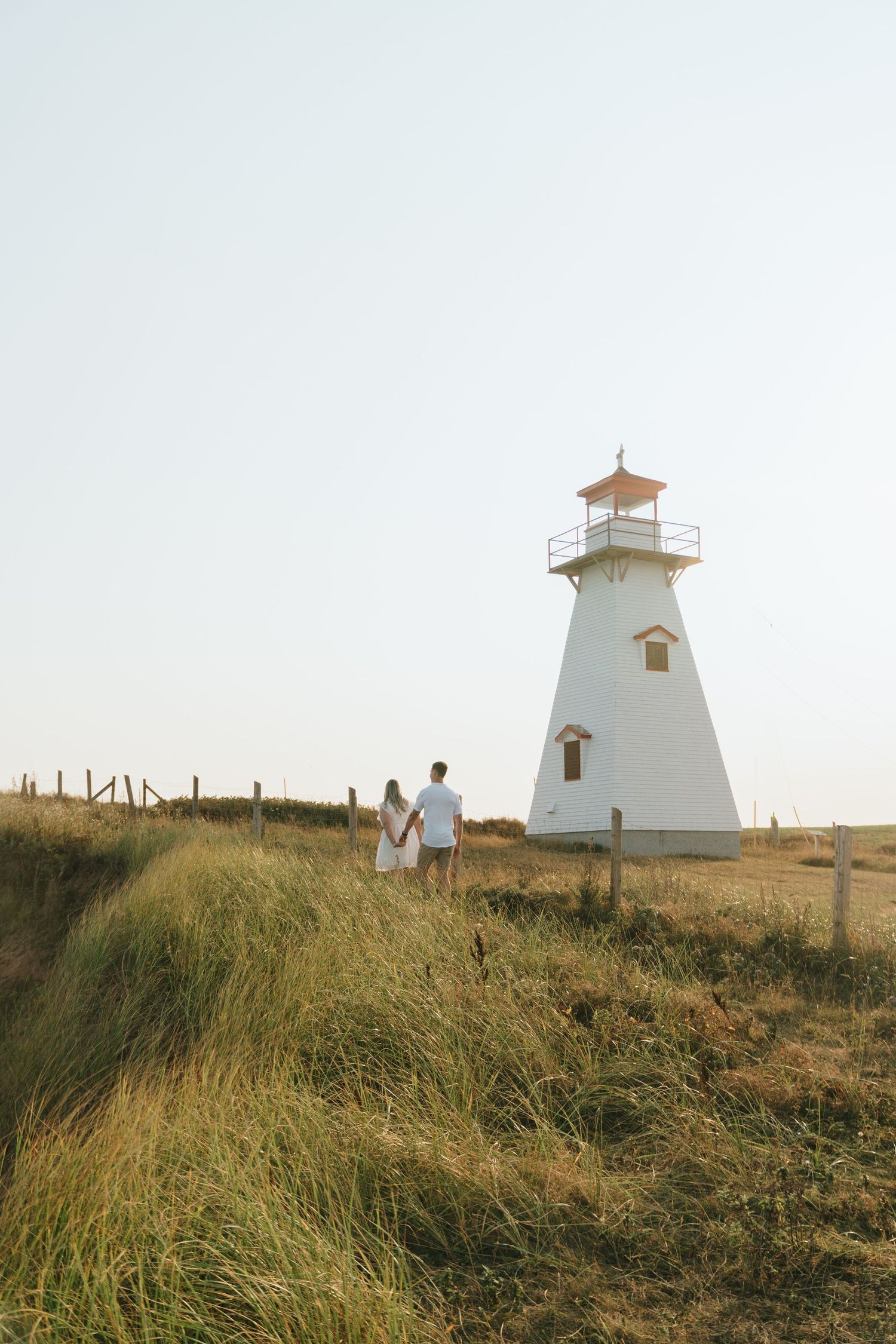 A white lighthouse stands tall on a grassy hill as two figures walk along the path at sunset.