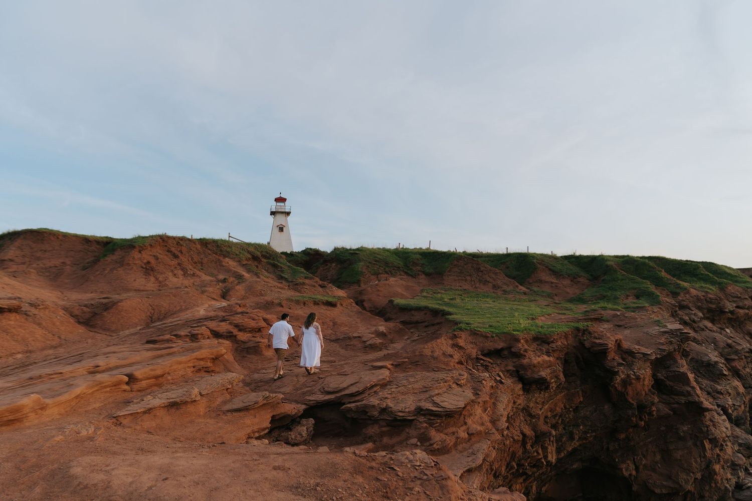 Two people in white clothing walk along red rocky cliffs near a lighthouse on the Hawaiian coast.