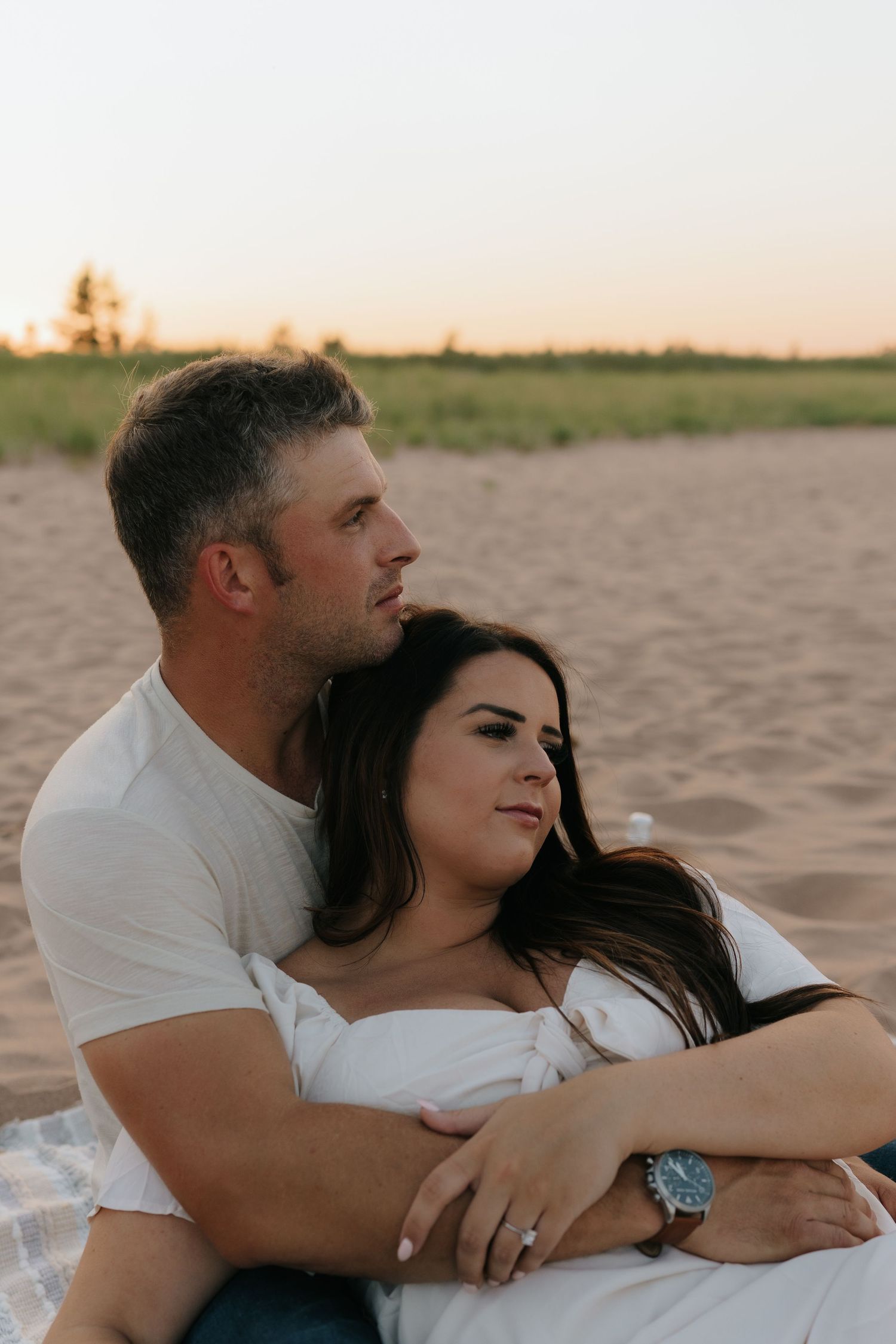 A couple embraces on the beach during sunset while sitting in the sand.