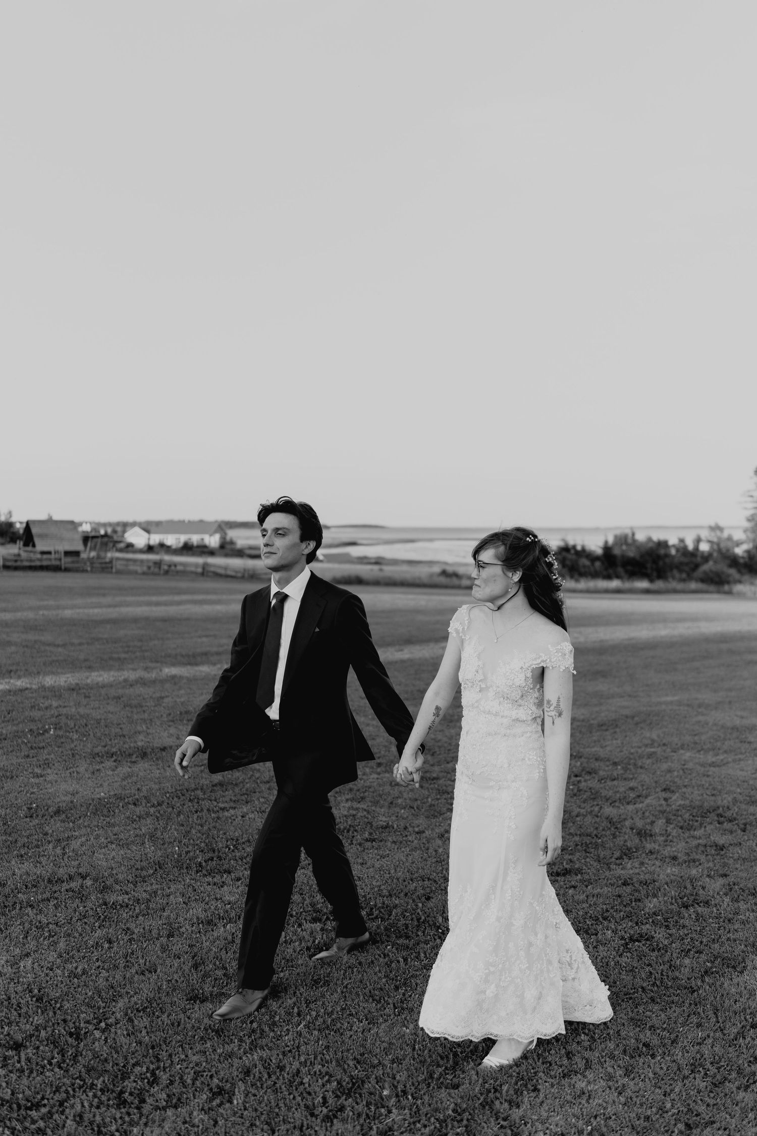 Black and white photo of newlyweds walking hand in hand across an open field.