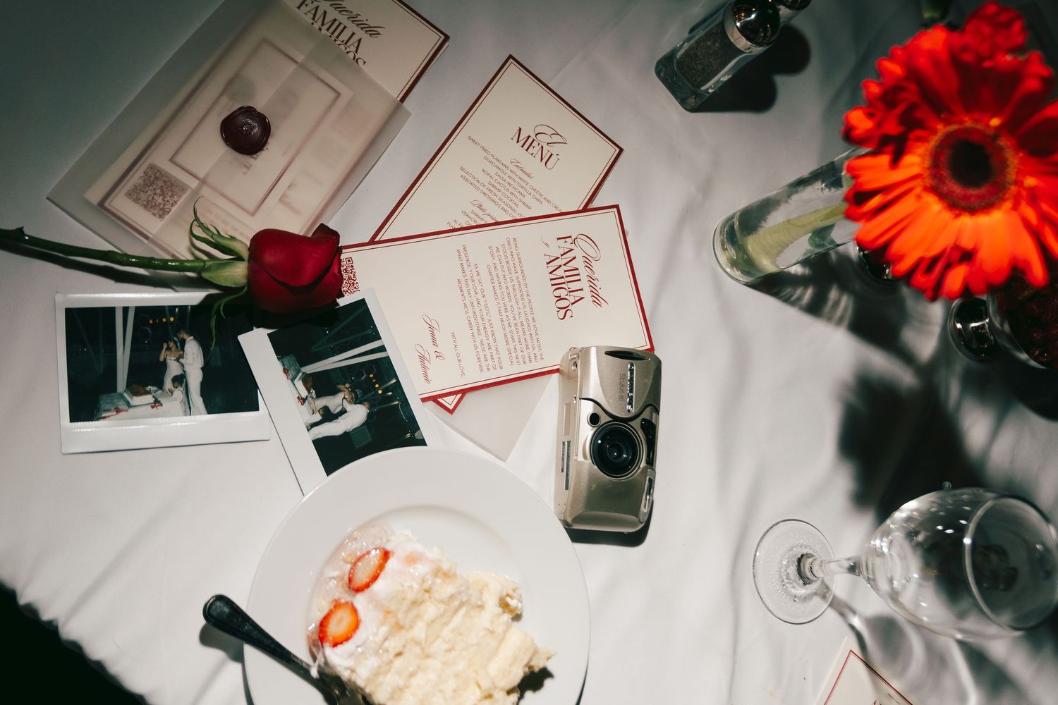 Wedding flat lay with red roses, polaroids, cake, cameras, menu cards, and champagne glasses on white tablecloth.