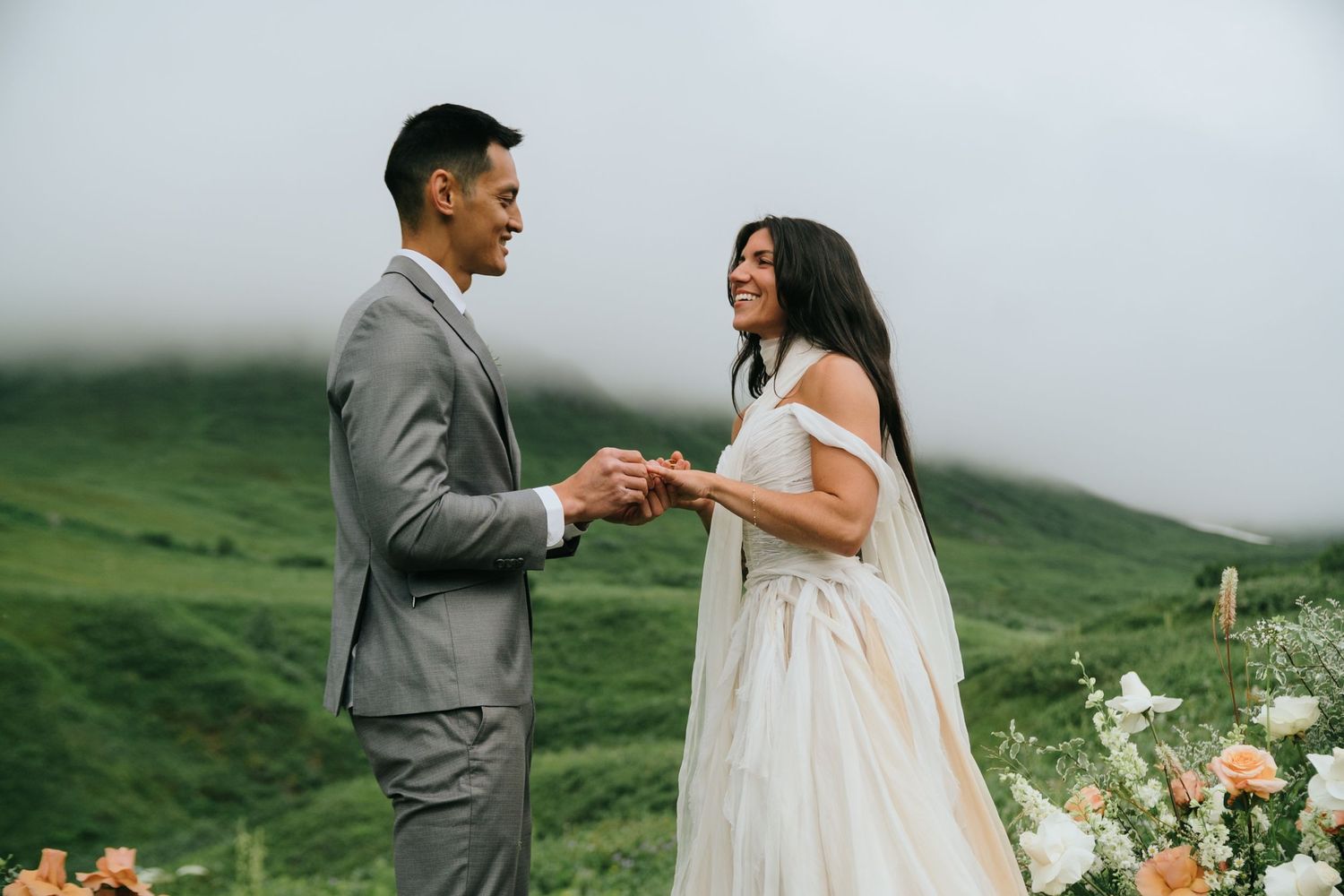 A couple shares an intimate moment during their wedding ceremony against a backdrop of lush green mountains in Alaska