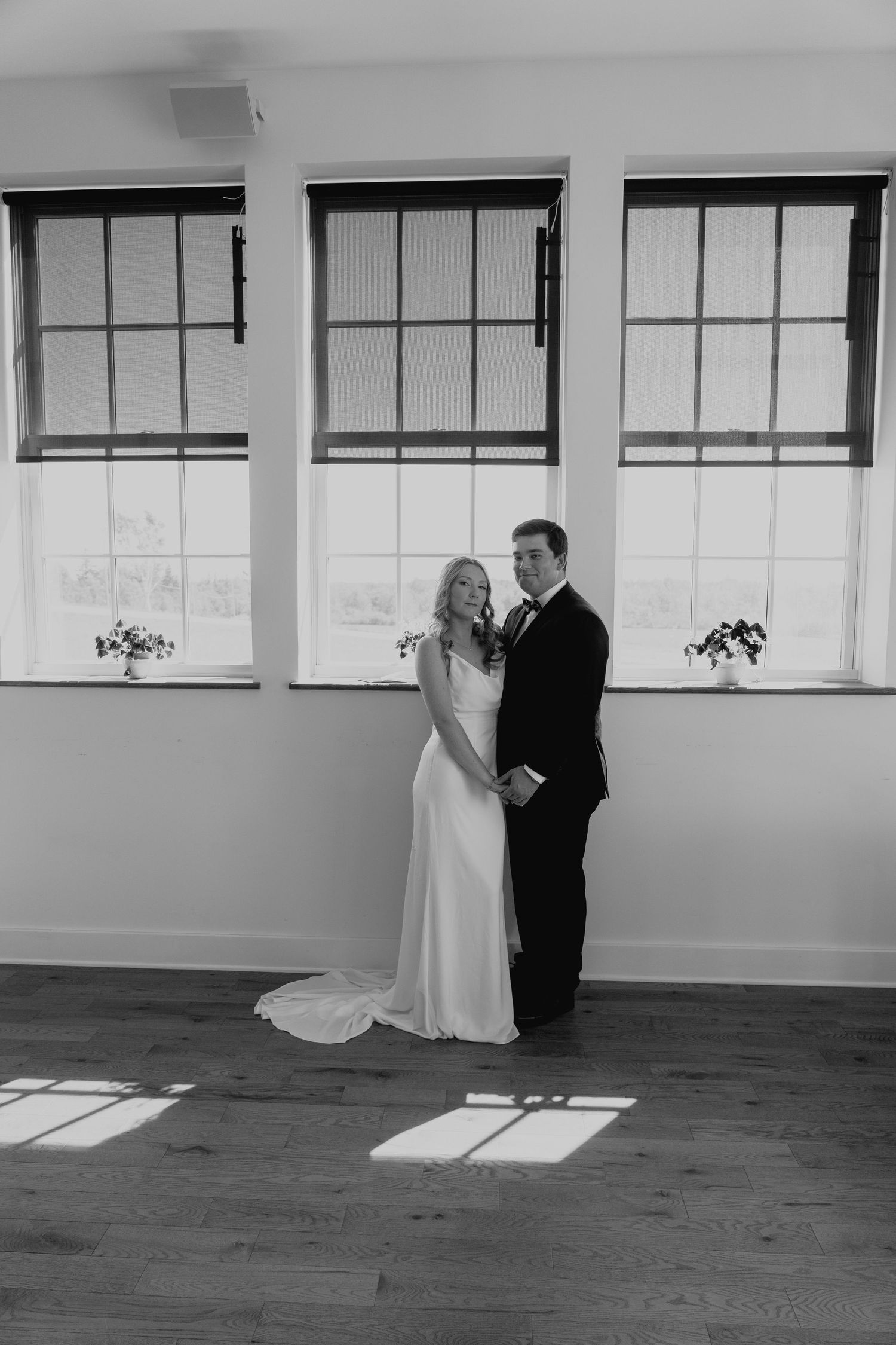 A couple in wedding attire poses in an empty room with large windows casting dramatic shadows.