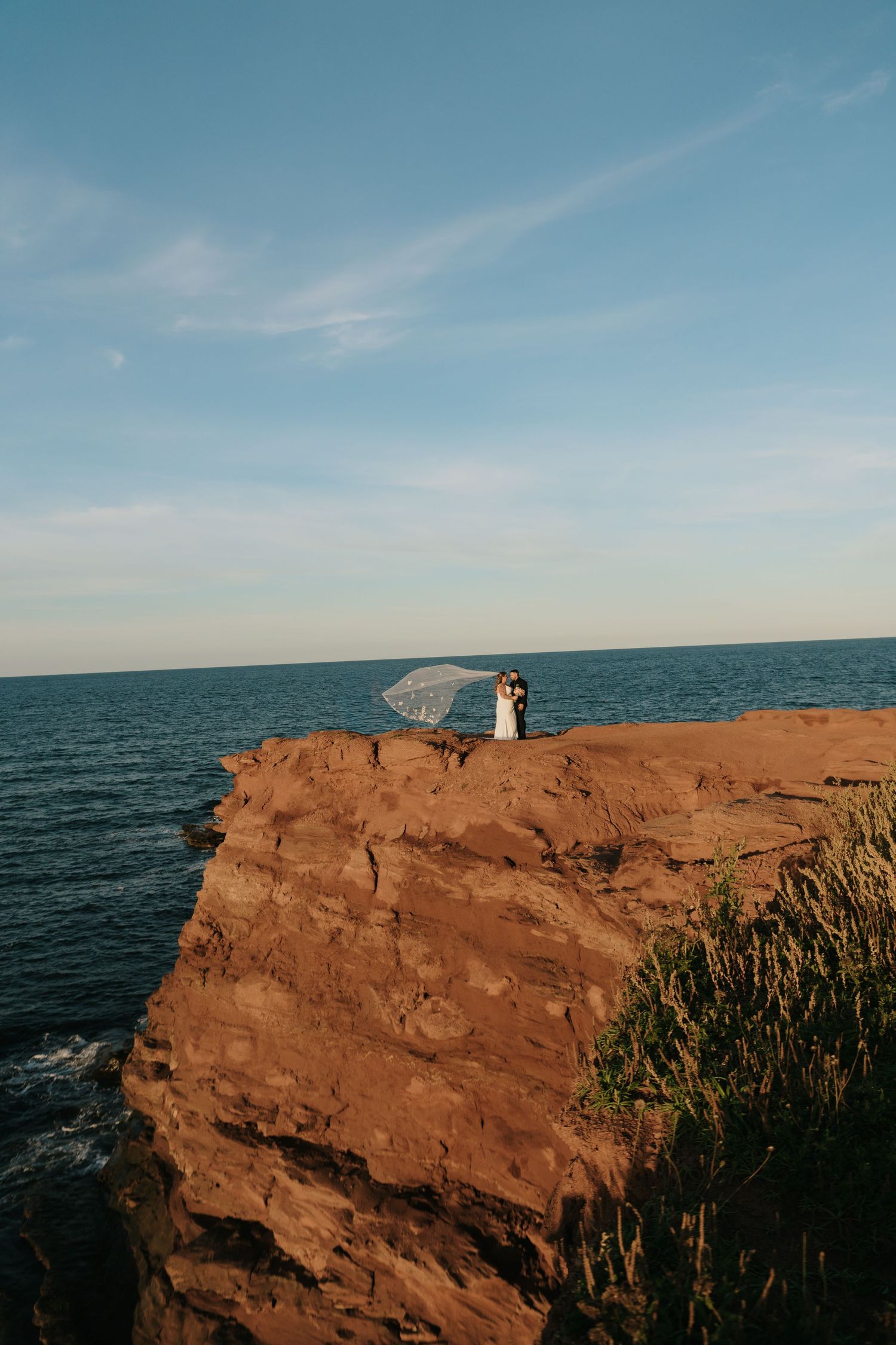 Bride and groom embrace on red cliffs overlooking the ocean, veil blowing in the wind under a blue sky.