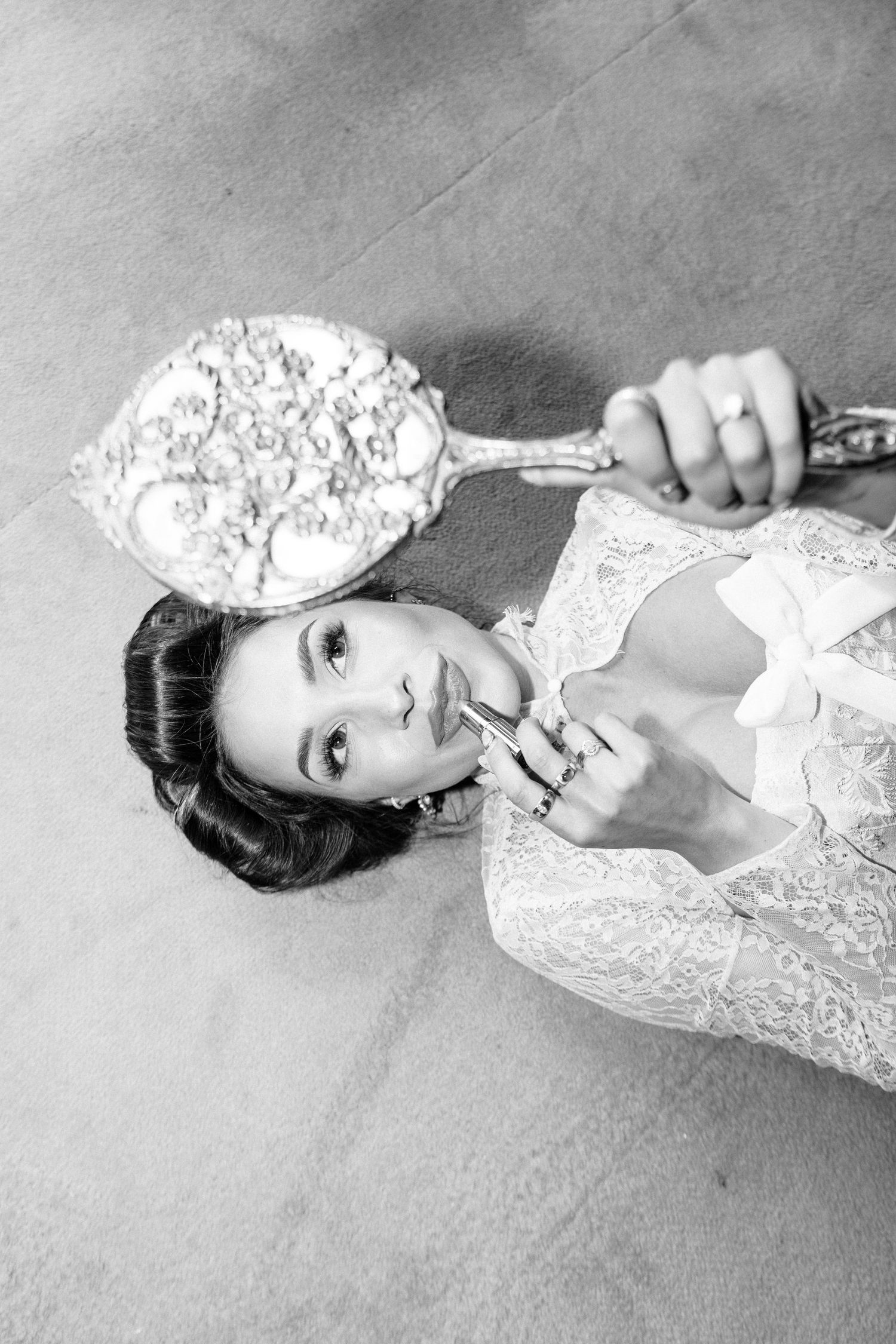 Bride lying on floor in lace robe applies lipstick while holding ornate vintage hand mirror, black and white photo.