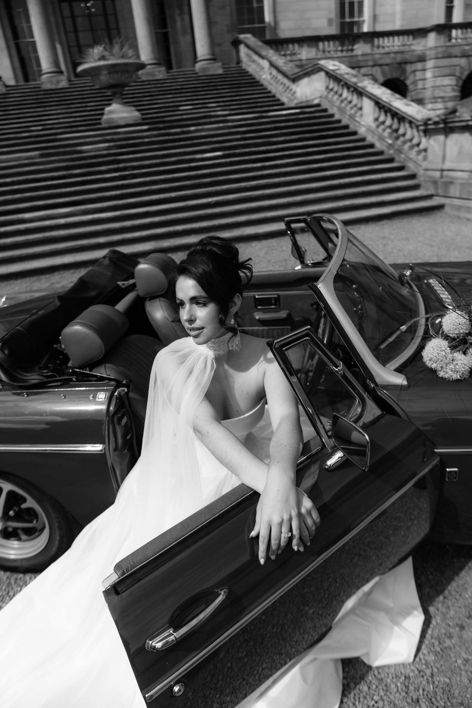 Black and white photo of a bride in a white gown leaning out of a vintage convertible car on grand stone steps.