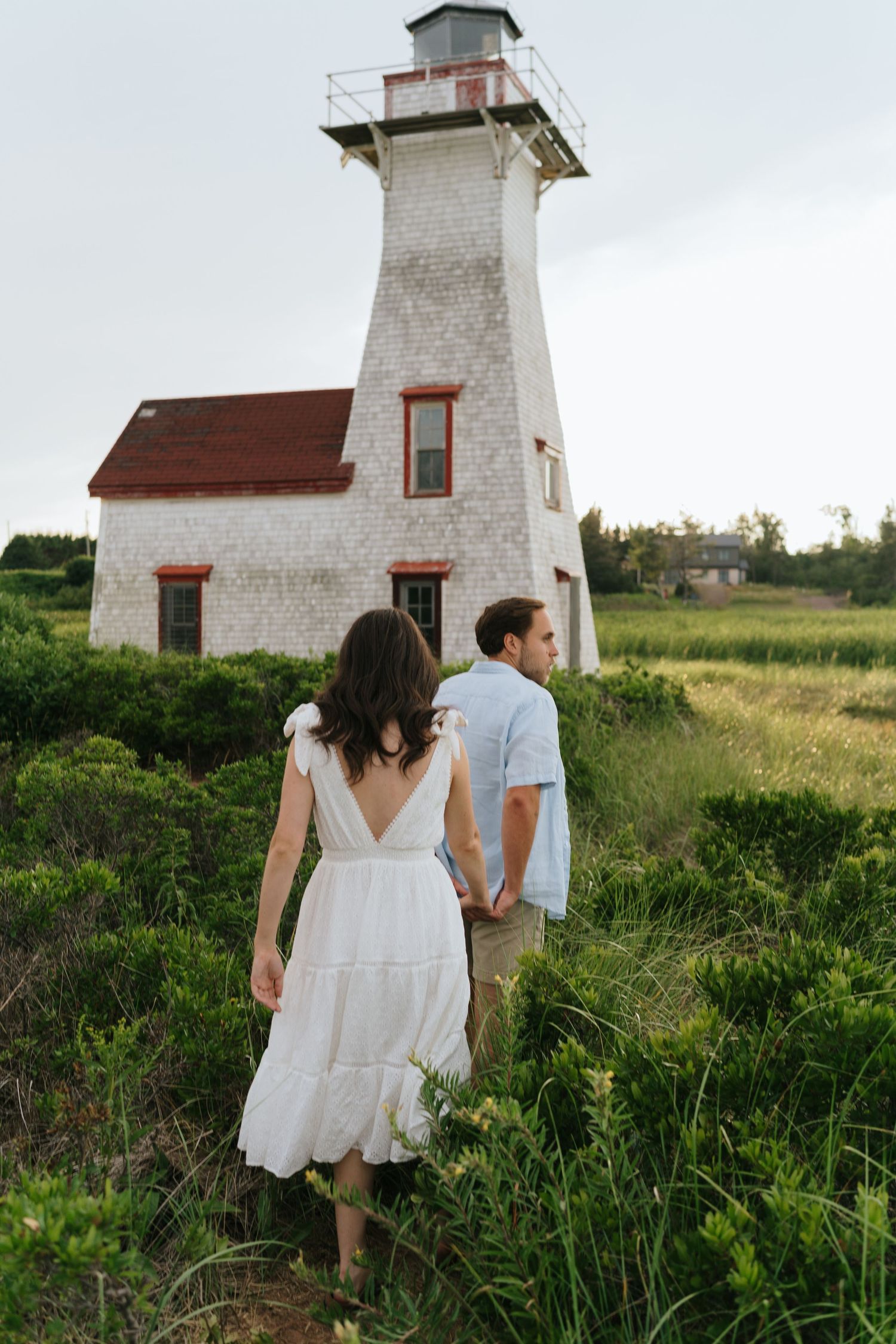 A couple walking hand in hand near a white lighthouse surrounded by green grass and wildflowers.