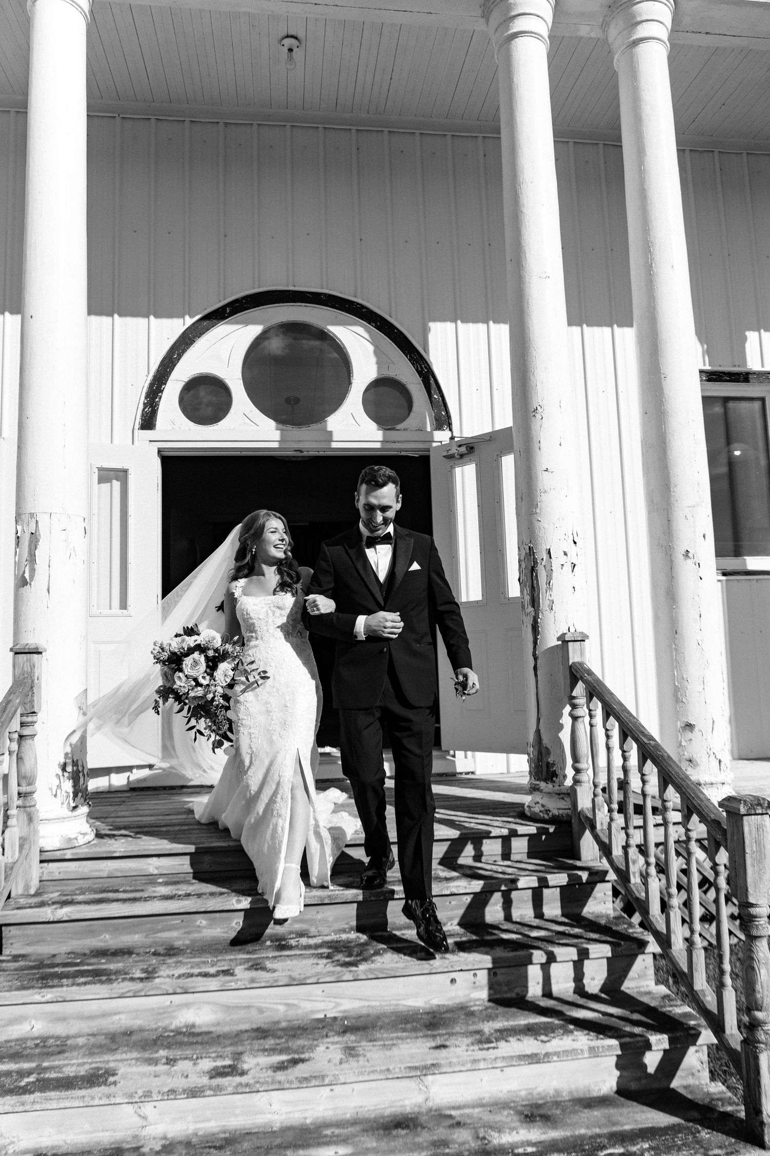 Black and white photo of a bride and groom descending church steps, bride holding bouquet, veil flowing in the wind.