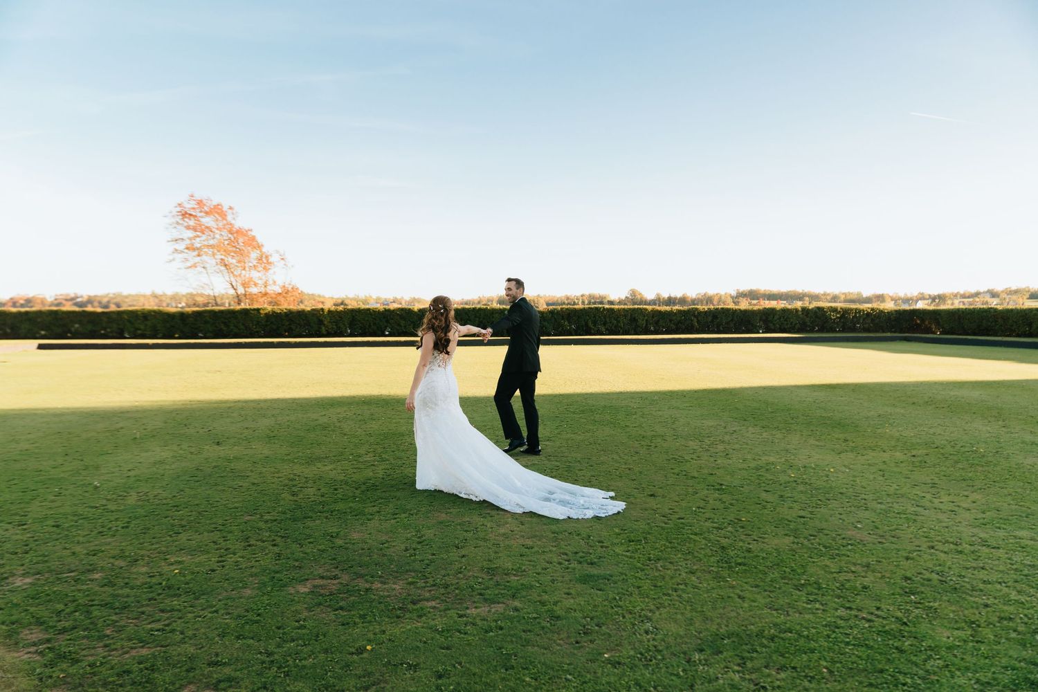 Bride and groom standing on lush green lawn at golden hour with autumn tree backdrop at outdoor wedding venue.