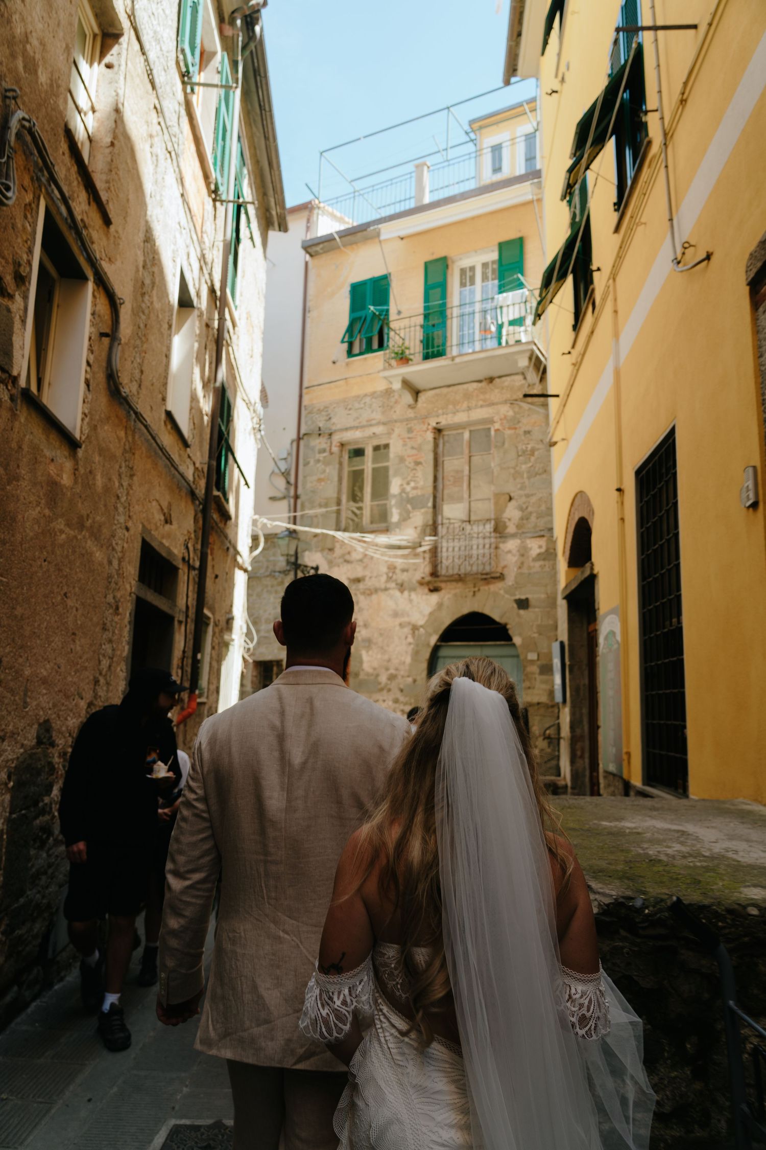 A couple in wedding attire walks through a charming narrow alley with yellow walls and green shutters in an Italian town.