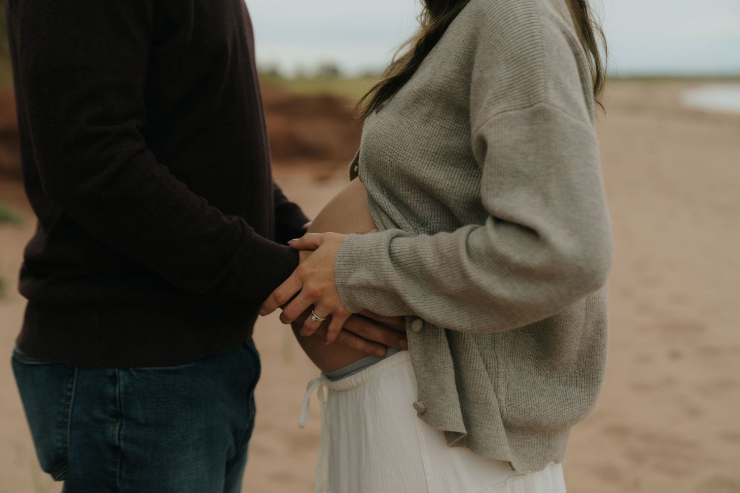 Couple holding hands tenderly on a beach, wearing casual sweaters and jeans, capturing a romantic engagement moment.