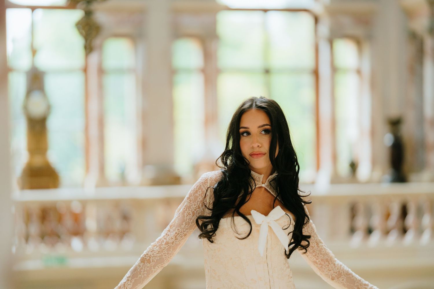 Elegant bride in white lace gown with long dark hair poses gracefully in ornate ballroom setting.