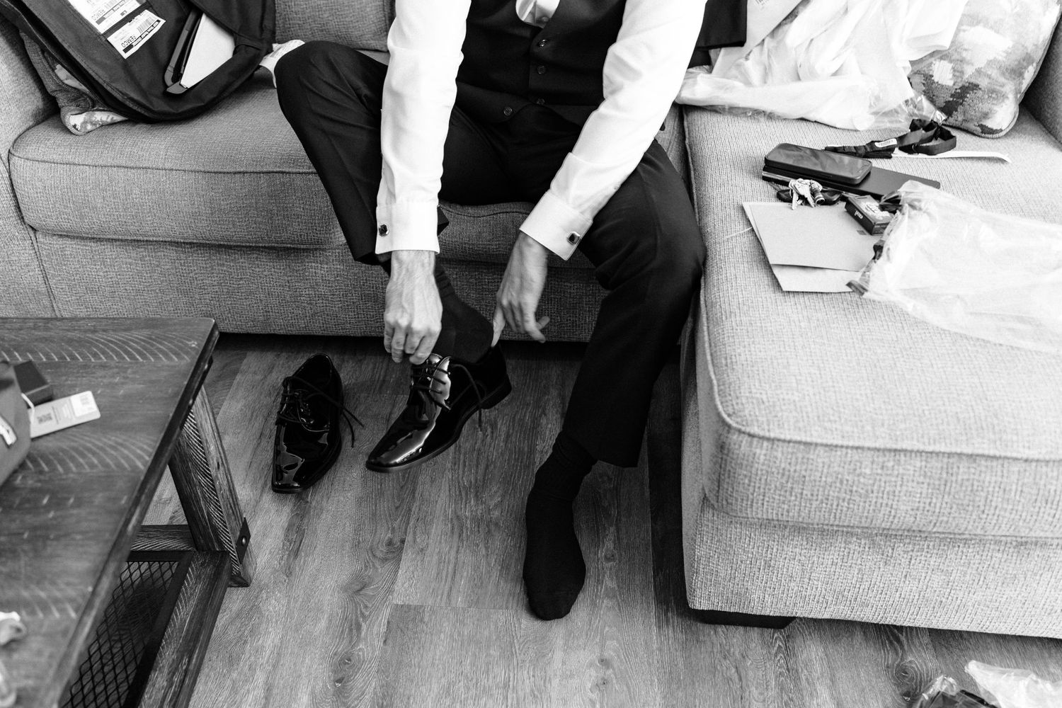 Black and white photo of a groom tying his dress shoes while seated on a couch during wedding preparations.