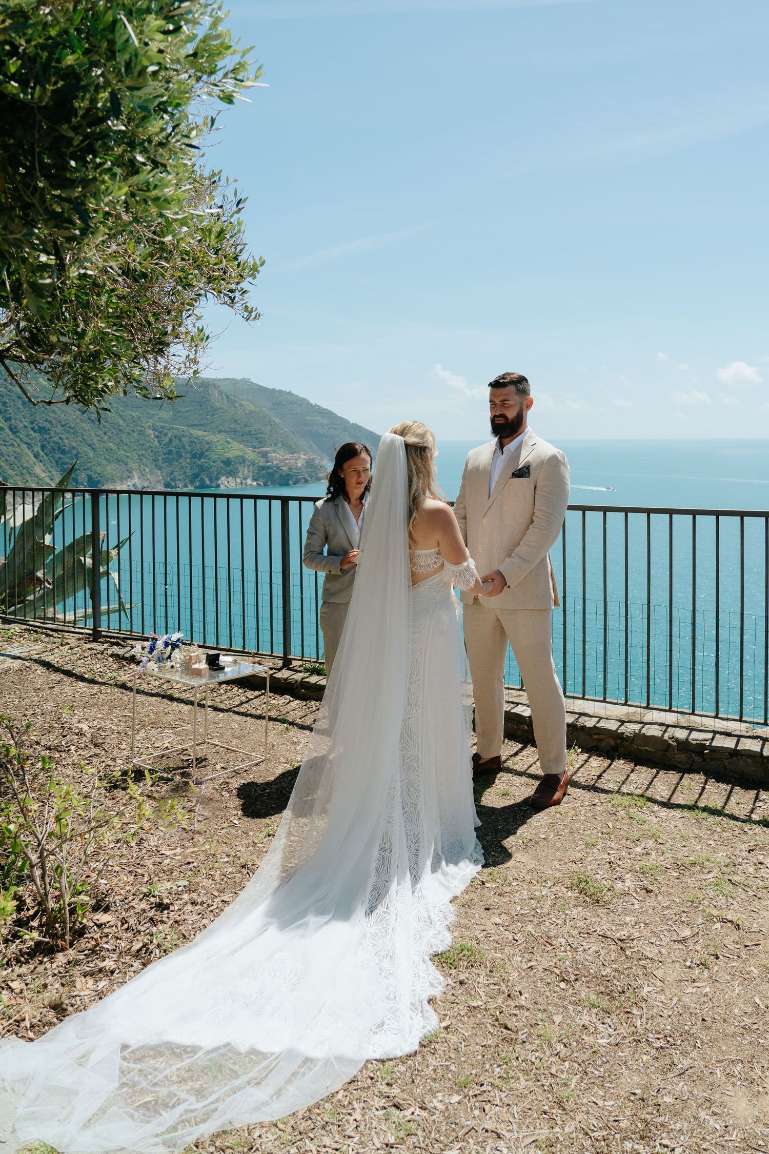 A wedding couple shares a romantic moment on a scenic coastal overlook with turquoise waters and mountains in the background.
