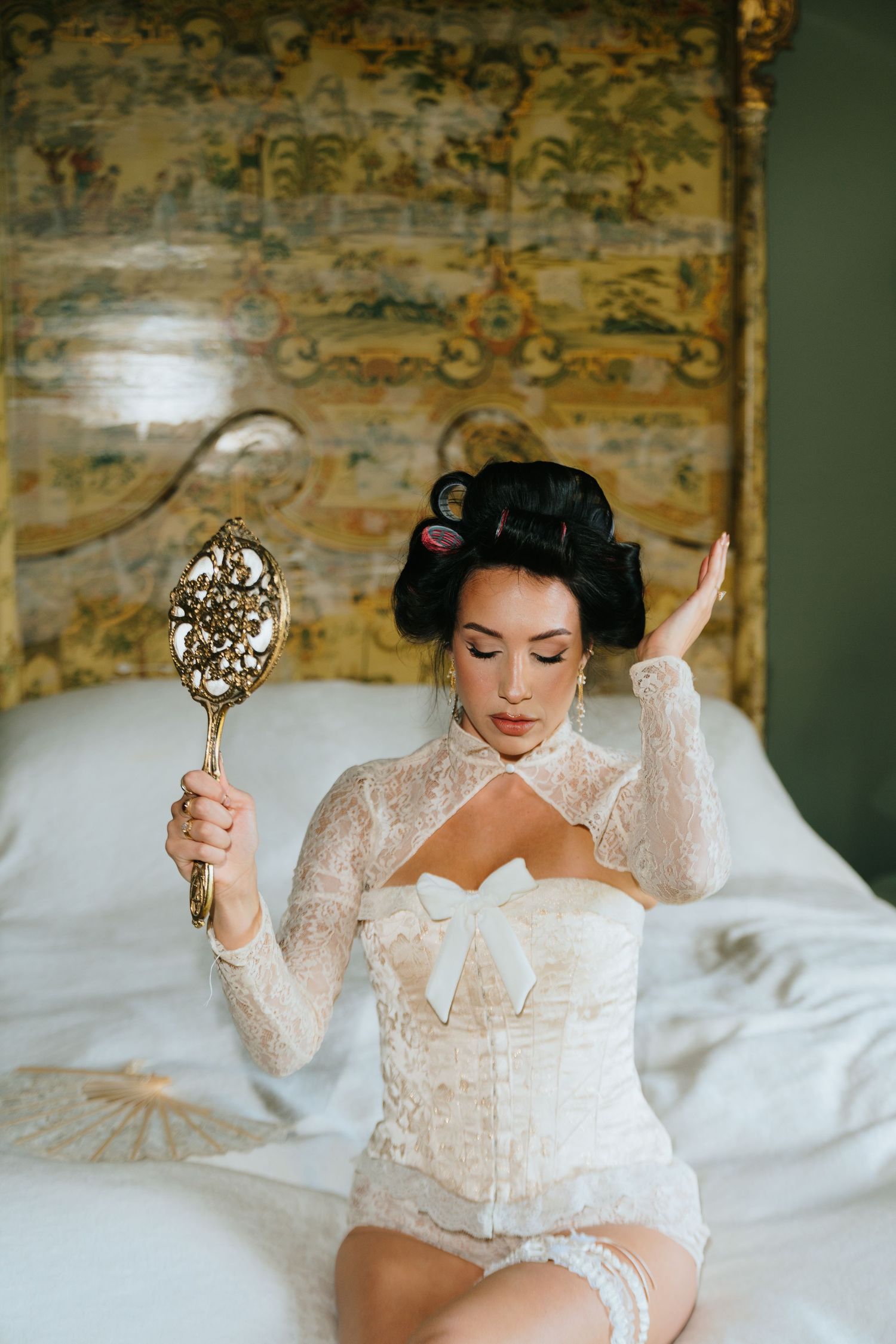Bride in white lace corset holding ornate gold hand mirror while sitting on bed.